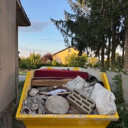 Patryk Kania - Żółty kontener wypełniony odpadami budowlanymi, meblami i workami, stojący na tle zieleni i zabudowań. Widoczne gruz, cegły, fragmenty mebli tapicerowanych.