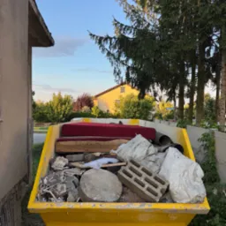 Żółty kontener wypełniony odpadami budowlanymi, meblami i workami, stojący na tle zieleni i zabudowań. Widoczne gruz, cegły, fragmenty mebli tapicerowanych.