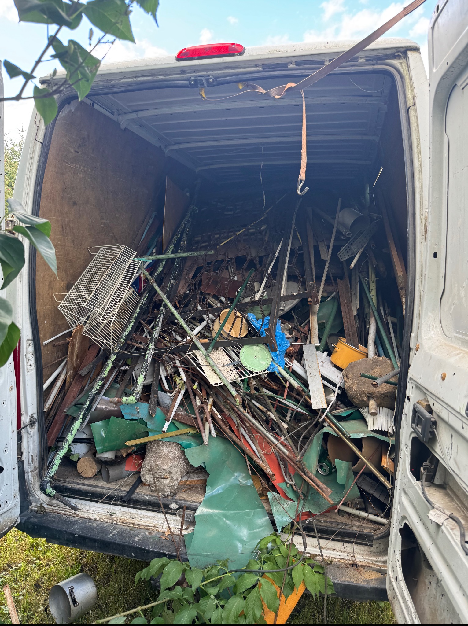 Wnętrze białego vana wypełnione różnorodnymi, nieposegregowanymi odpadami metalowymi, elementami konstrukcyjnymi i przedmiotami gospodarstwa domowego.