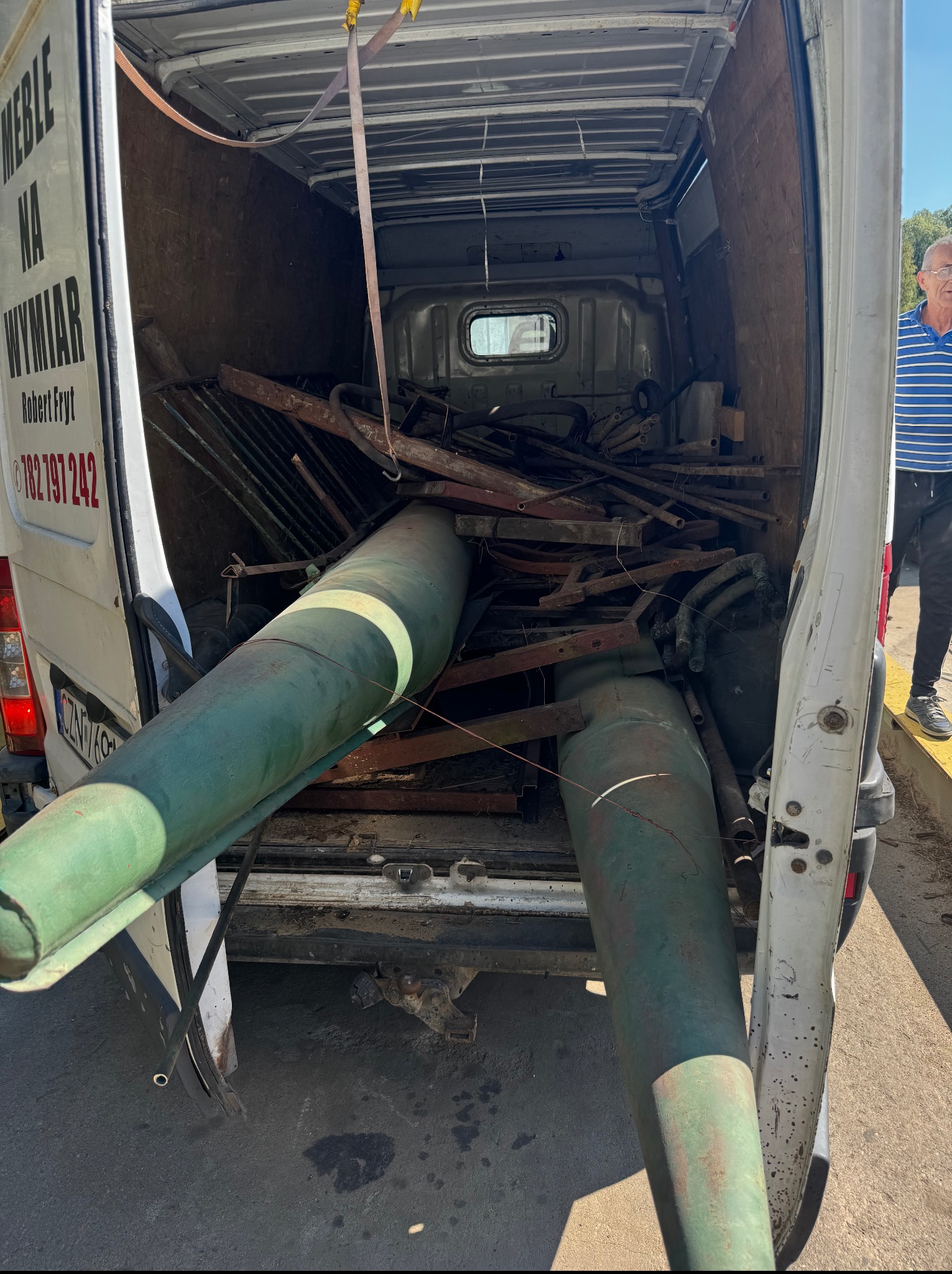 Wnętrze busa załadowane elementami metalowymi, rurami i konstrukcjami. Widoczne także butle gazowe. Częściowo widoczny bok busa z reklamą firmy.