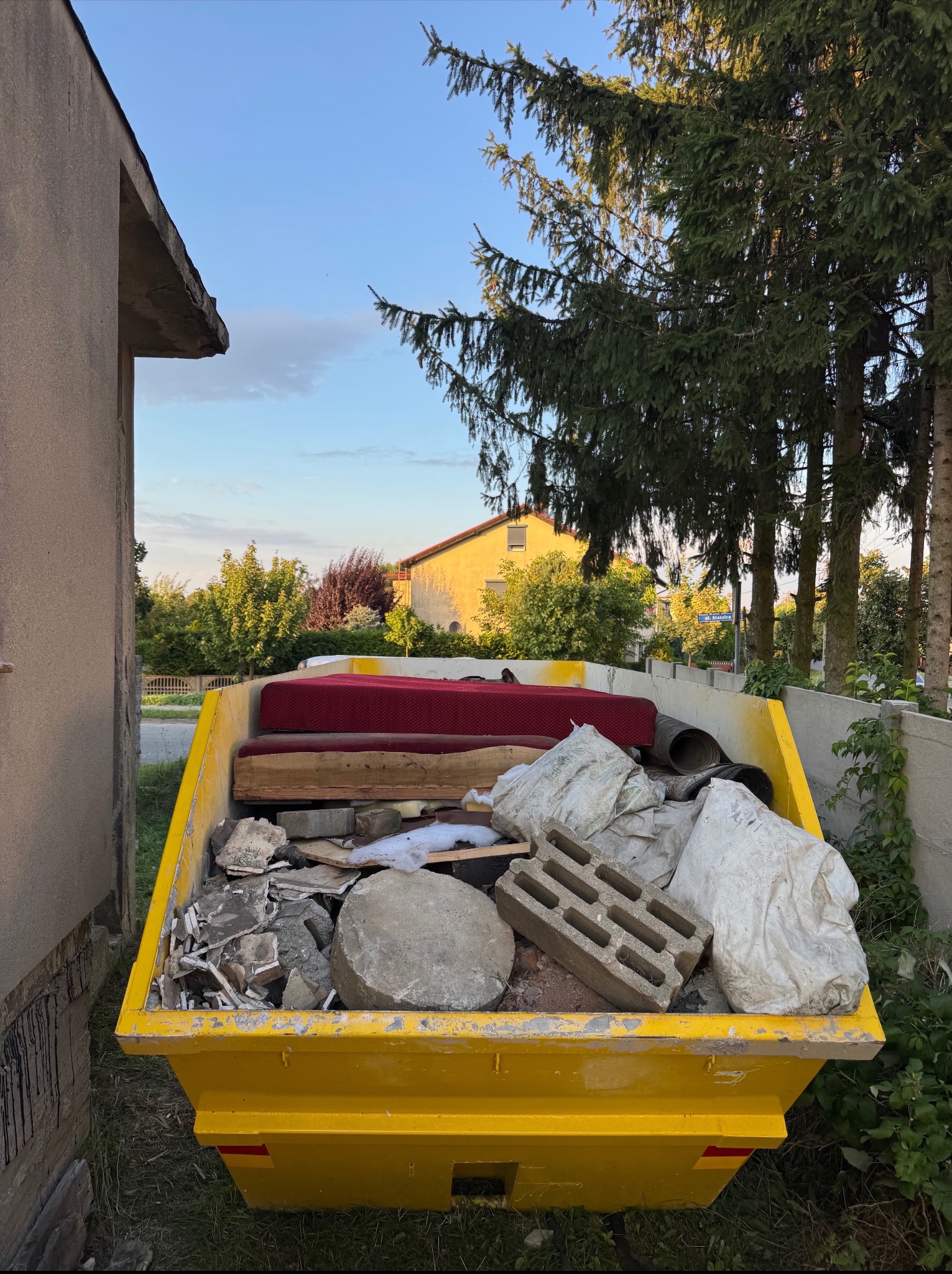 Żółty kontener wypełniony odpadami budowlanymi, meblami i workami, stojący na tle zieleni i zabudowań. Widoczne gruz, cegły, fragmenty mebli tapicerowanych.