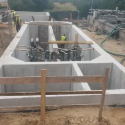 Budowa żelbetowej konstrukcji zbiornika retencyjnego z widocznym zbrojeniem i pracownikami w żółtych kamizelkach na placu budowy.