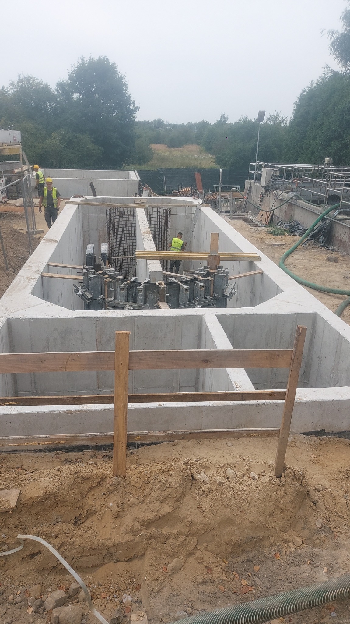 Budowa żelbetowej konstrukcji zbiornika retencyjnego z widocznym zbrojeniem i pracownikami w żółtych kamizelkach na placu budowy.