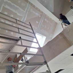 Trockenbau Team - Montaż płyt gipsowo-kartonowych na poddaszu ze świetlikiem. Widoczne rusztowania, pracownicy podczas pracy i częściowo wyszpachlowane łączenia płyt.