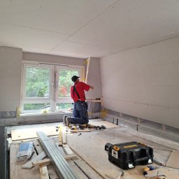 Trockenbau Team - Pracownik w czerwonej koszulce montuje płytę gipsową przy oknie. Widoczne narzędzia, skrzynka DeWalt i częściowo zamontowane płyty na ścianach i suficie. Wnętrze w trakcie prac.