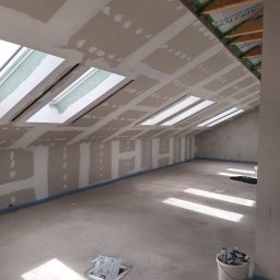 Trockenbau Team - Poddasze z oknami dachowymi w trakcie montażu płyt gipsowo-kartonowych. Widoczne łączenia płyt i szpachlowanie. Na podłodze wiadro z narzędziami.