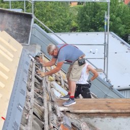 Trockenbau Team - Demontaż starego pokrycia dachowego przez dwóch mężczyzn w okularach, jeden z nich w szelkach, na tle zieleni i rusztowania. Widoczne narzędzia i fragmenty dachówki.