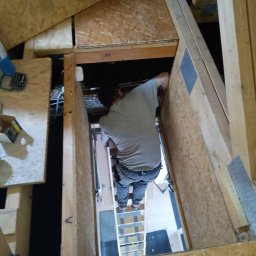 Trockenbau Team - Pracownik montuje płytę OSB w przestrzeni poddasza, widoczna drabina i elementy konstrukcyjne dachu. Ujęcie z góry, podkreślające trudność dostępu.