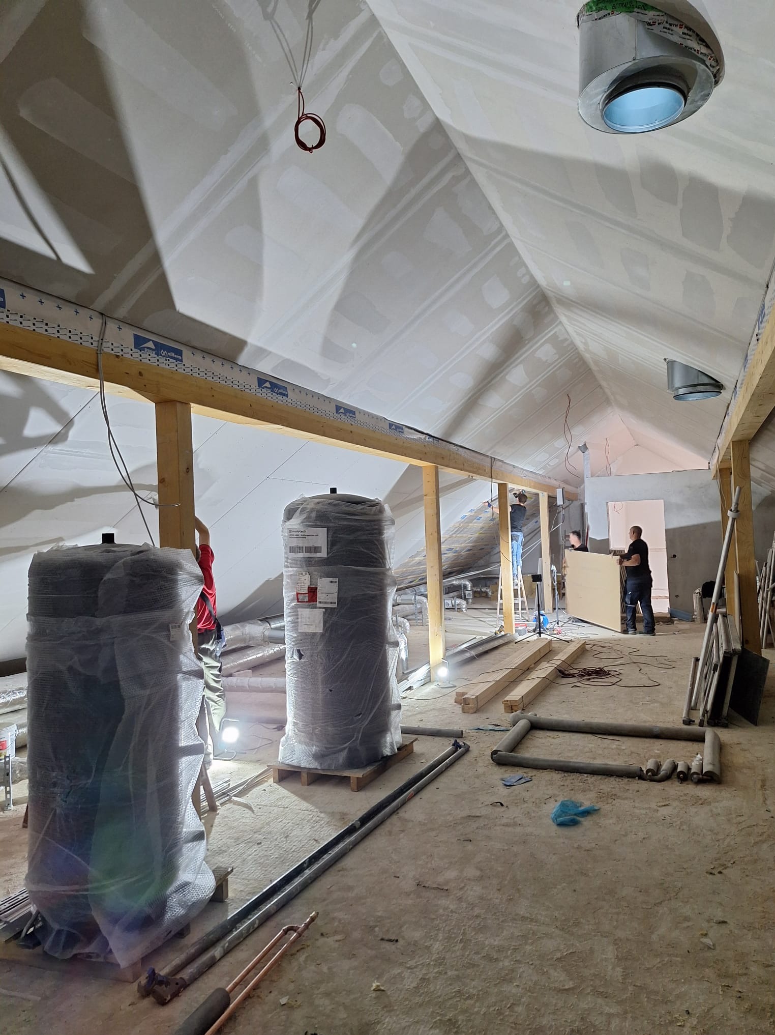 Poddasze w trakcie adaptacji: widoczne płyty gipsowe na skosach, drewniana konstrukcja, ocieplenie w folii i pracownicy montujący ściankę działową w głębi pomieszczenia.