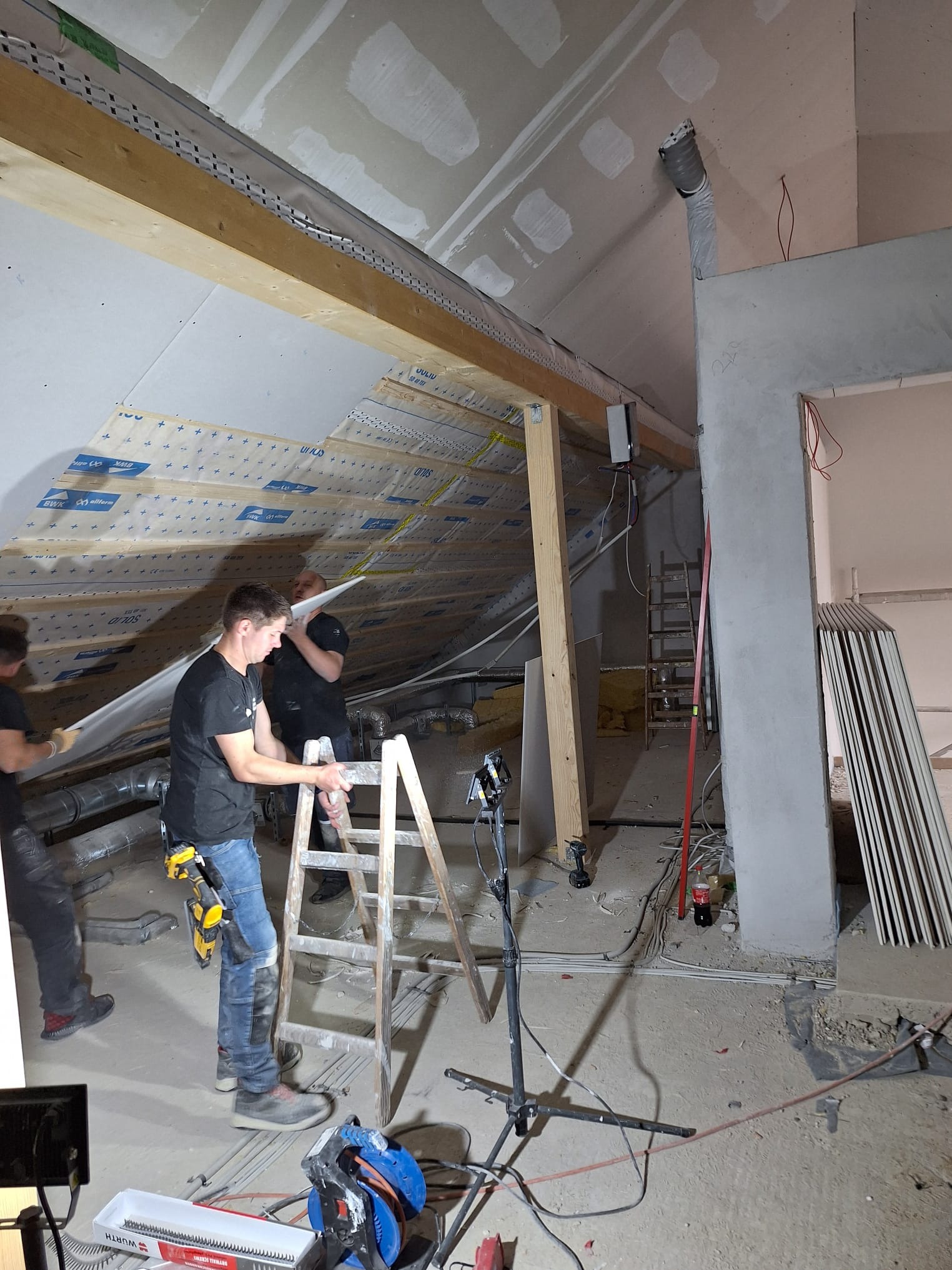 Montaż płyt kartonowo-gipsowych na poddaszu. Widoczni trzej pracownicy na drabinie, narzędzia i materiały budowlane. Ujęcie z dołu, surowe wykończenie.