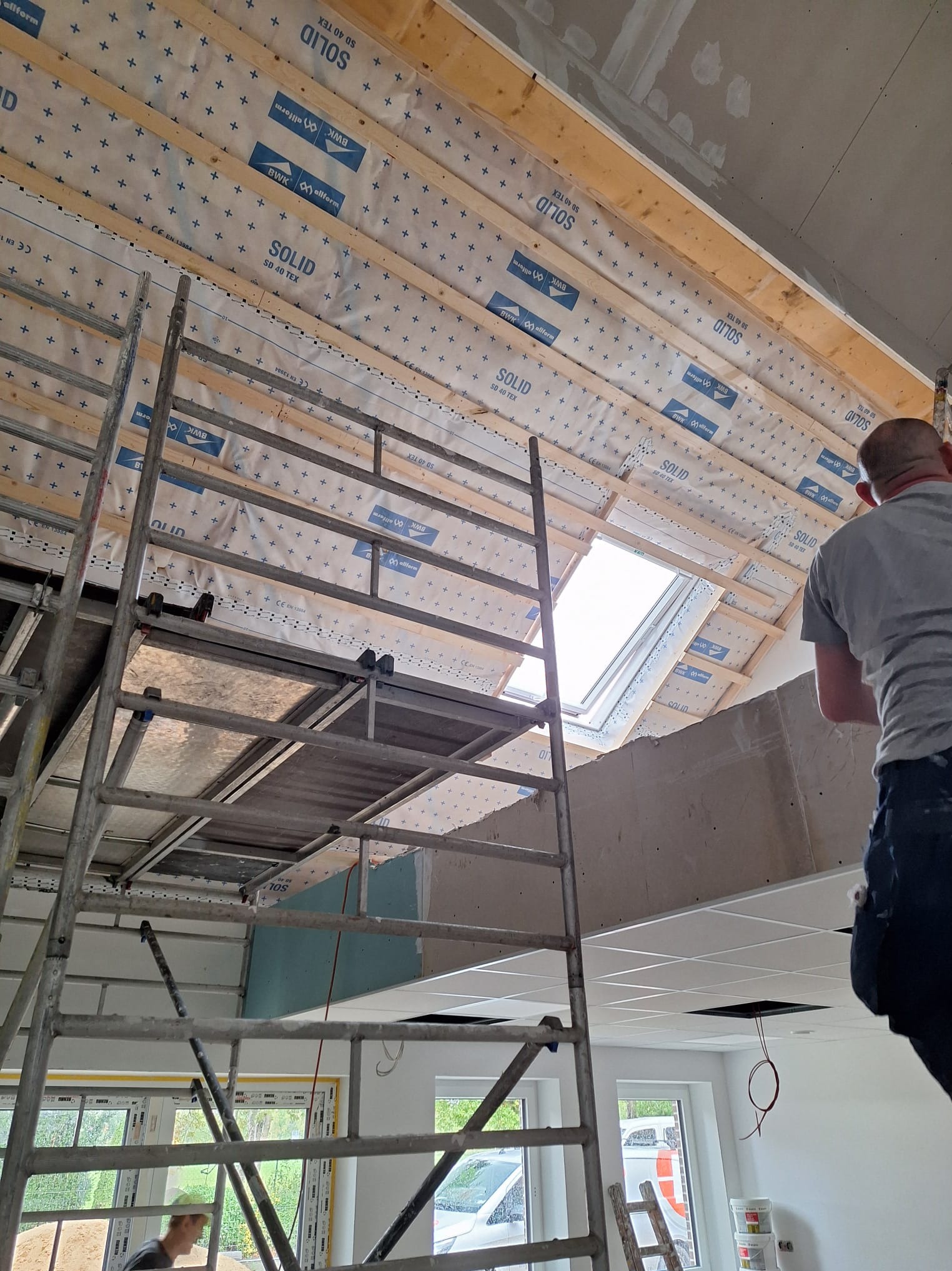 Ocieplenie poddasza wełną mineralną widoczne spod rusztowania. Skosy dachu z oknem połaciowym, folia paroizolacyjna z nadrukiem, fragment płyty karton-gips.