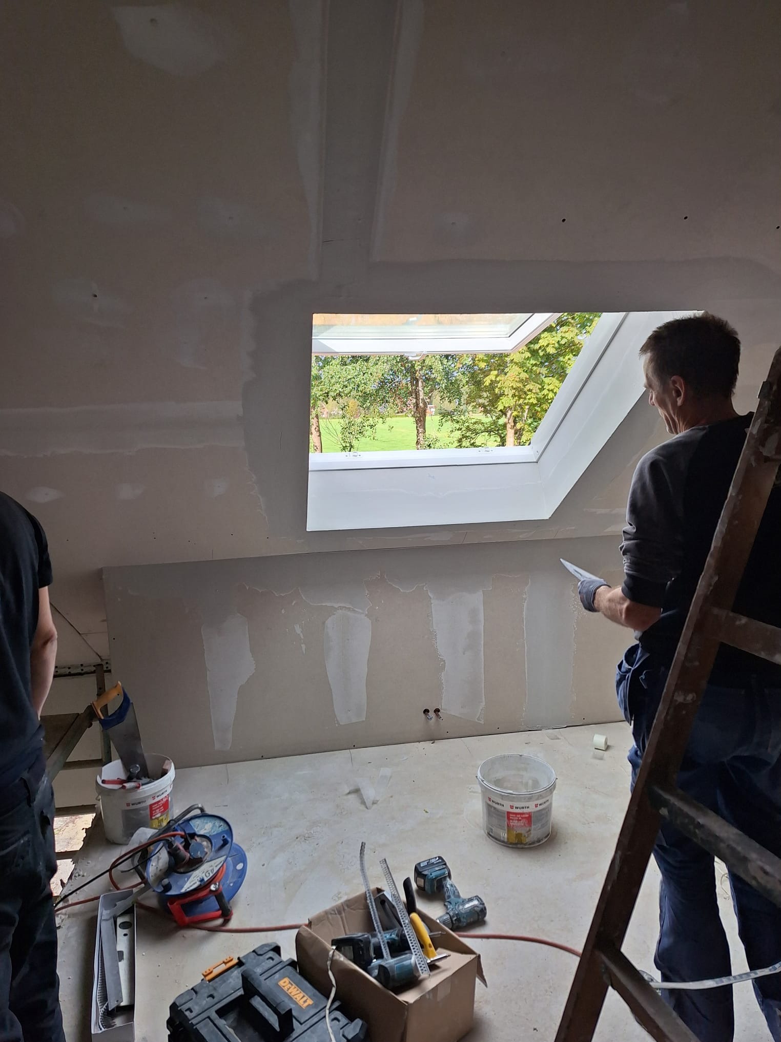 Poddasze z oknem dachowym w trakcie szpachlowania. Widoczne narzędzia, drabina i fragment sylwetki pracownika. Ściany przygotowane pod gładzie.
