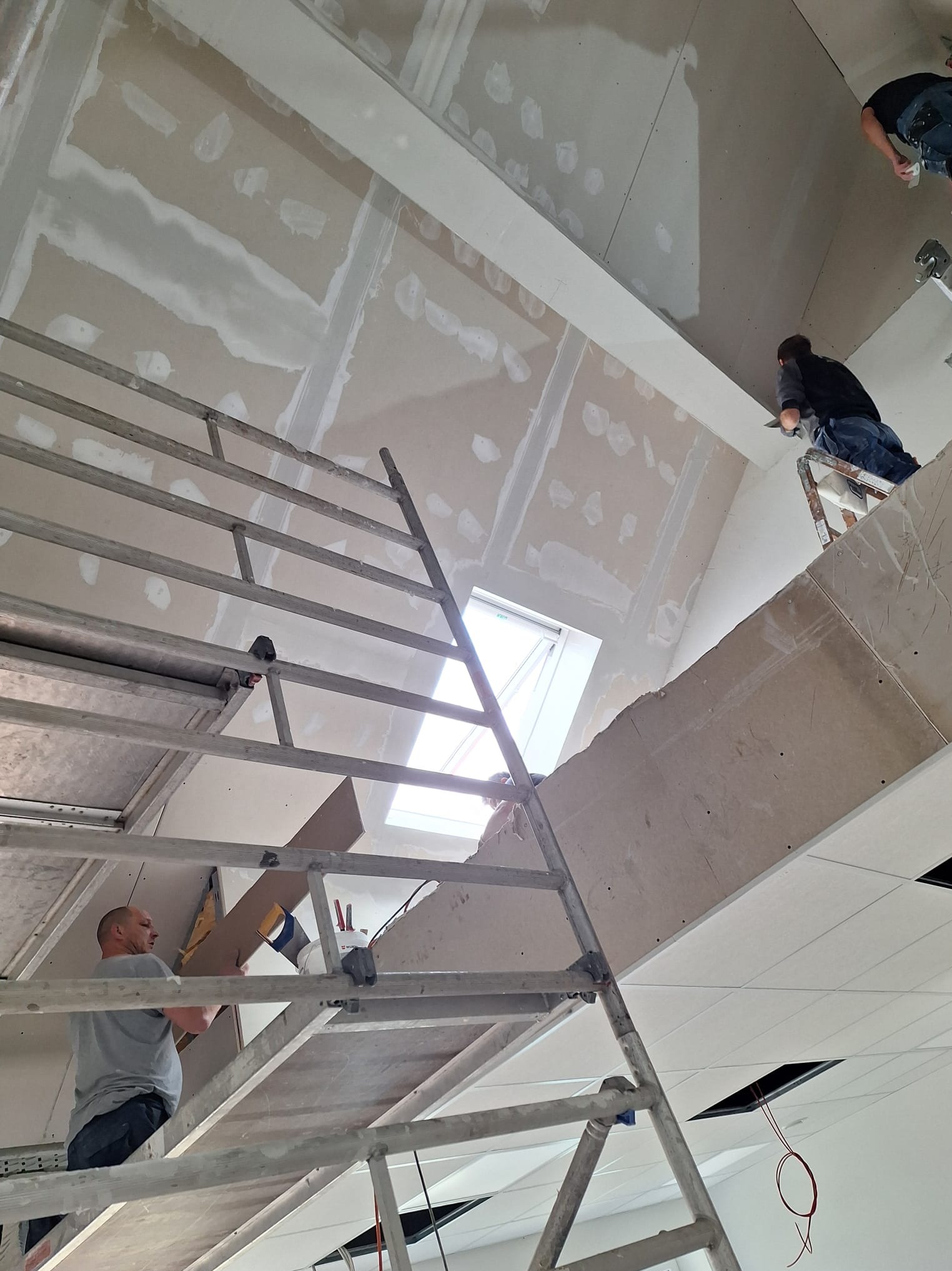 Montaż płyt gipsowo-kartonowych na poddaszu ze świetlikiem. Widoczne rusztowania, pracownicy podczas pracy i częściowo wyszpachlowane łączenia płyt.