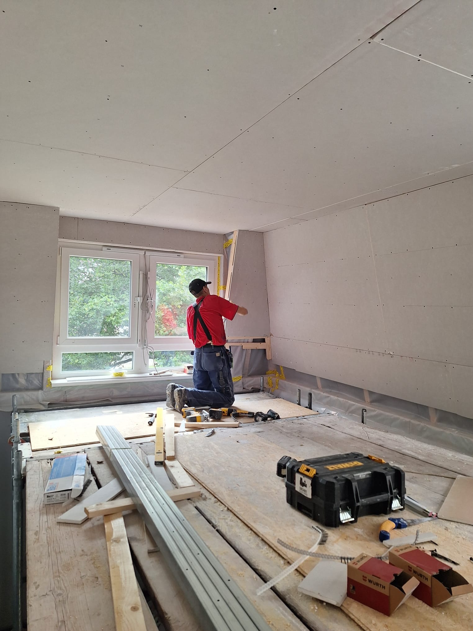 Pracownik w czerwonej koszulce montuje płytę gipsową przy oknie. Widoczne narzędzia, skrzynka DeWalt i częściowo zamontowane płyty na ścianach i suficie. Wnętrze w trakcie prac.