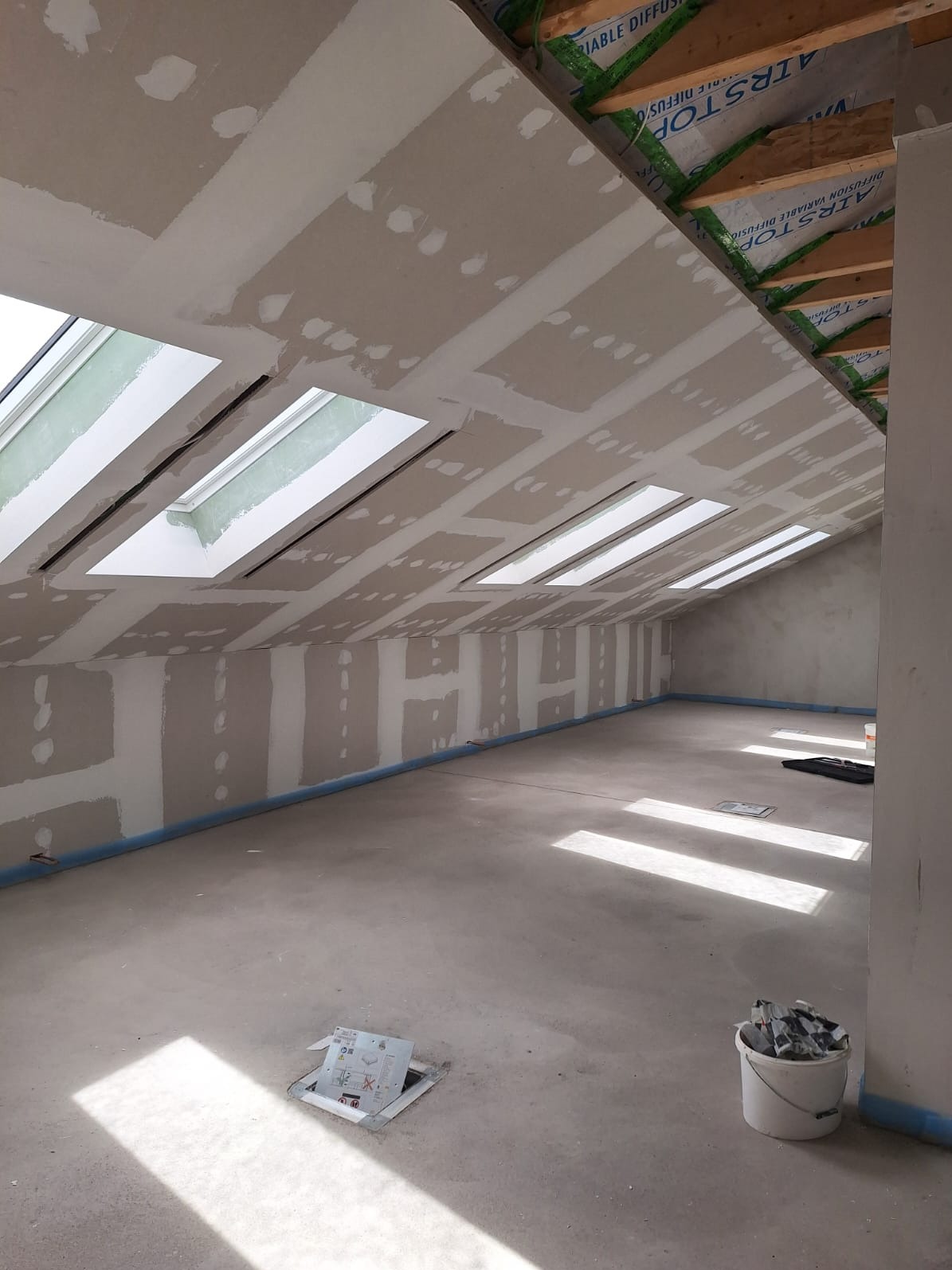 Poddasze z oknami dachowymi w trakcie montażu płyt gipsowo-kartonowych. Widoczne łączenia płyt i szpachlowanie. Na podłodze wiadro z narzędziami.