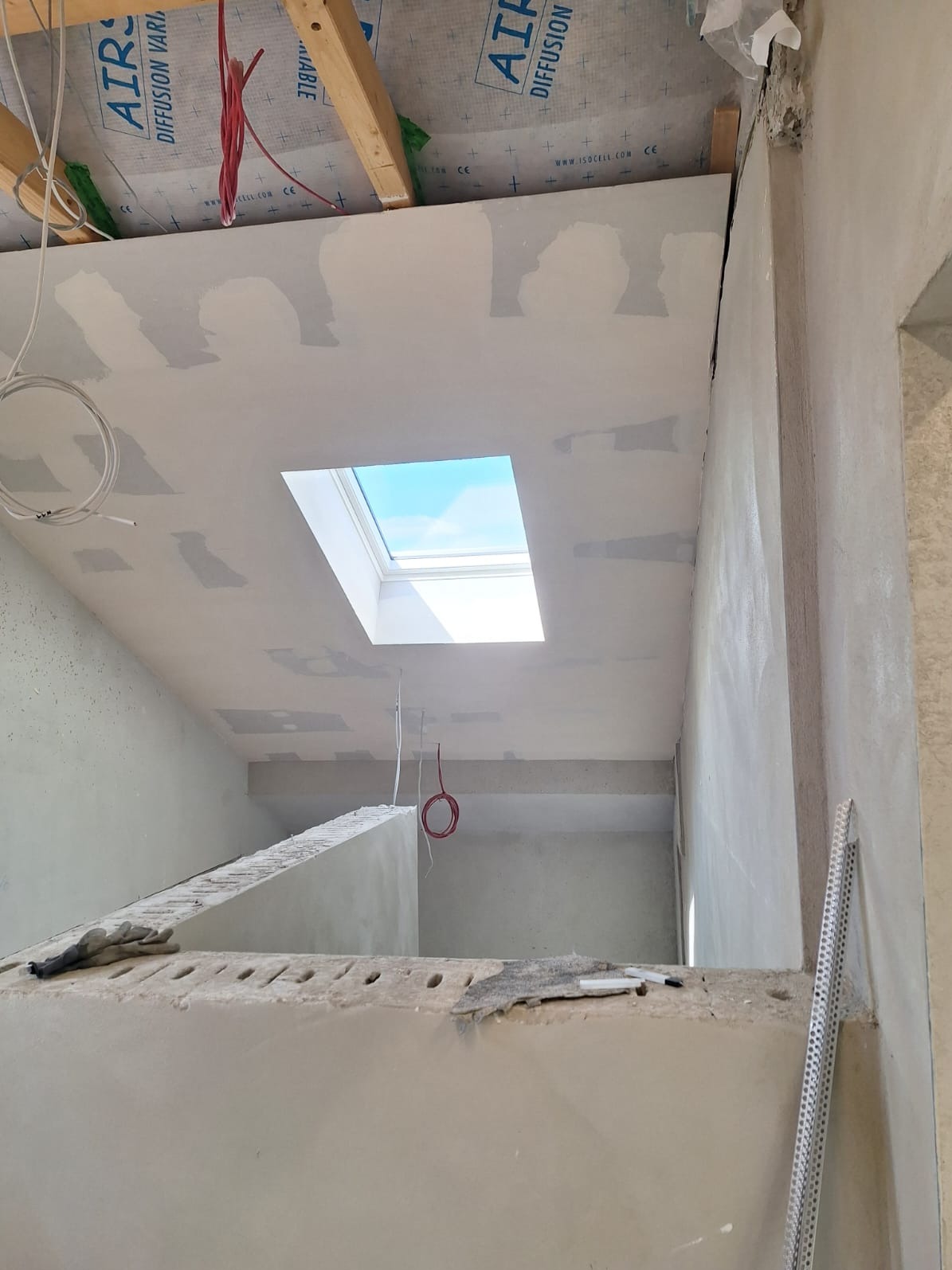 Poddasze z oknem dachowym w trakcie prac wykończeniowych: szpachlowanie sufitu i ścian. Widoczne przewody elektryczne i fragment konstrukcji dachu z membraną.