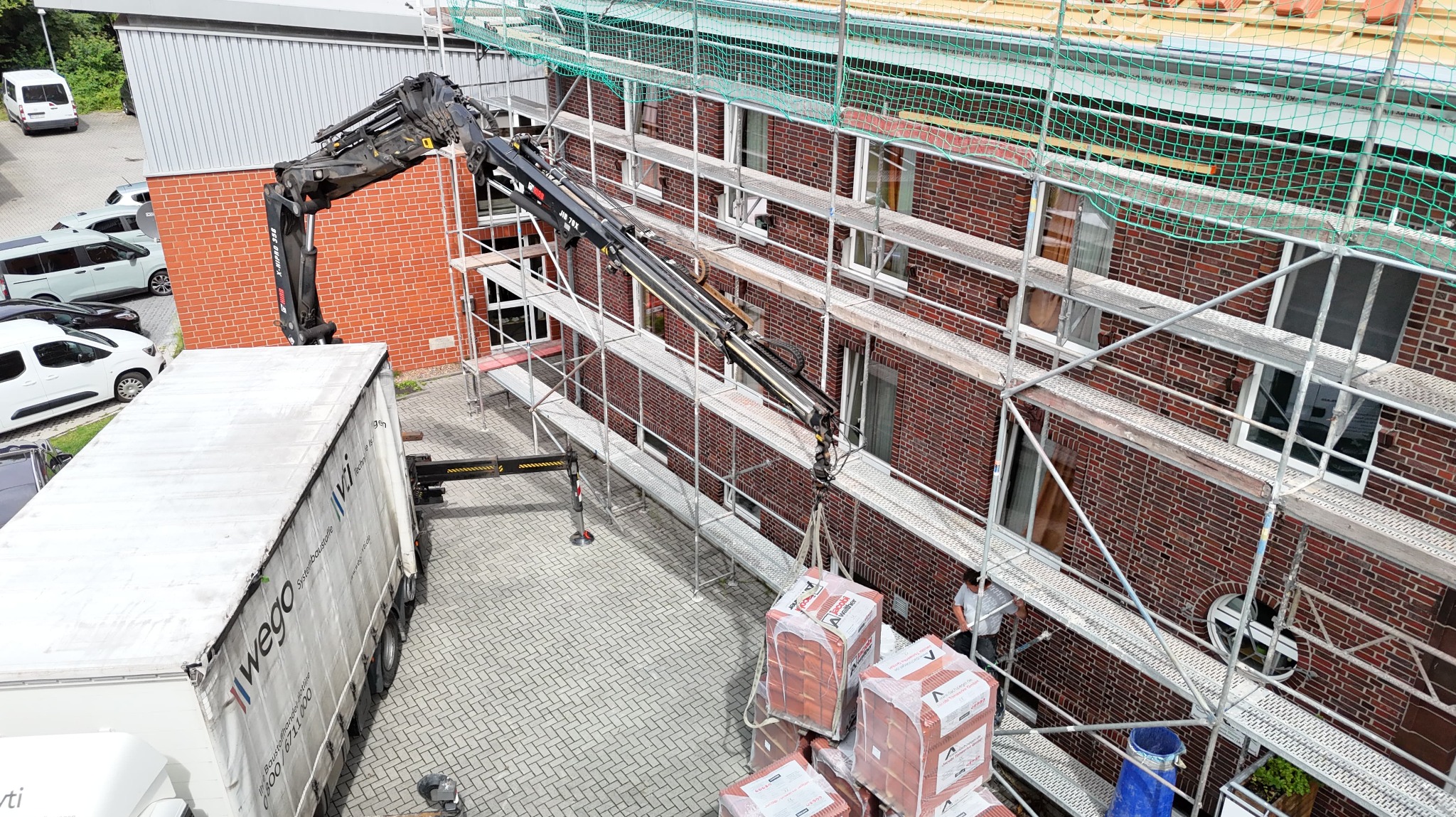 Ceglana elewacja budynku w rusztowaniach, z dźwigiem podającym materiały. Widok z góry na plac budowy z paletami cegieł i ciężarówką dostawczą.