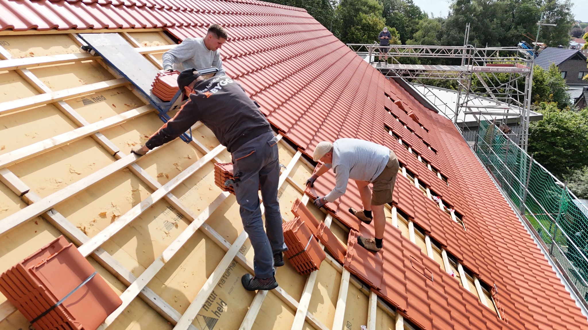 Montaż czerwonej dachówki na drewnianej konstrukcji dachu przez trzech pracowników. Widoczne rusztowanie i zabezpieczenia. Ujęcie z dołu, w słoneczny dzień.