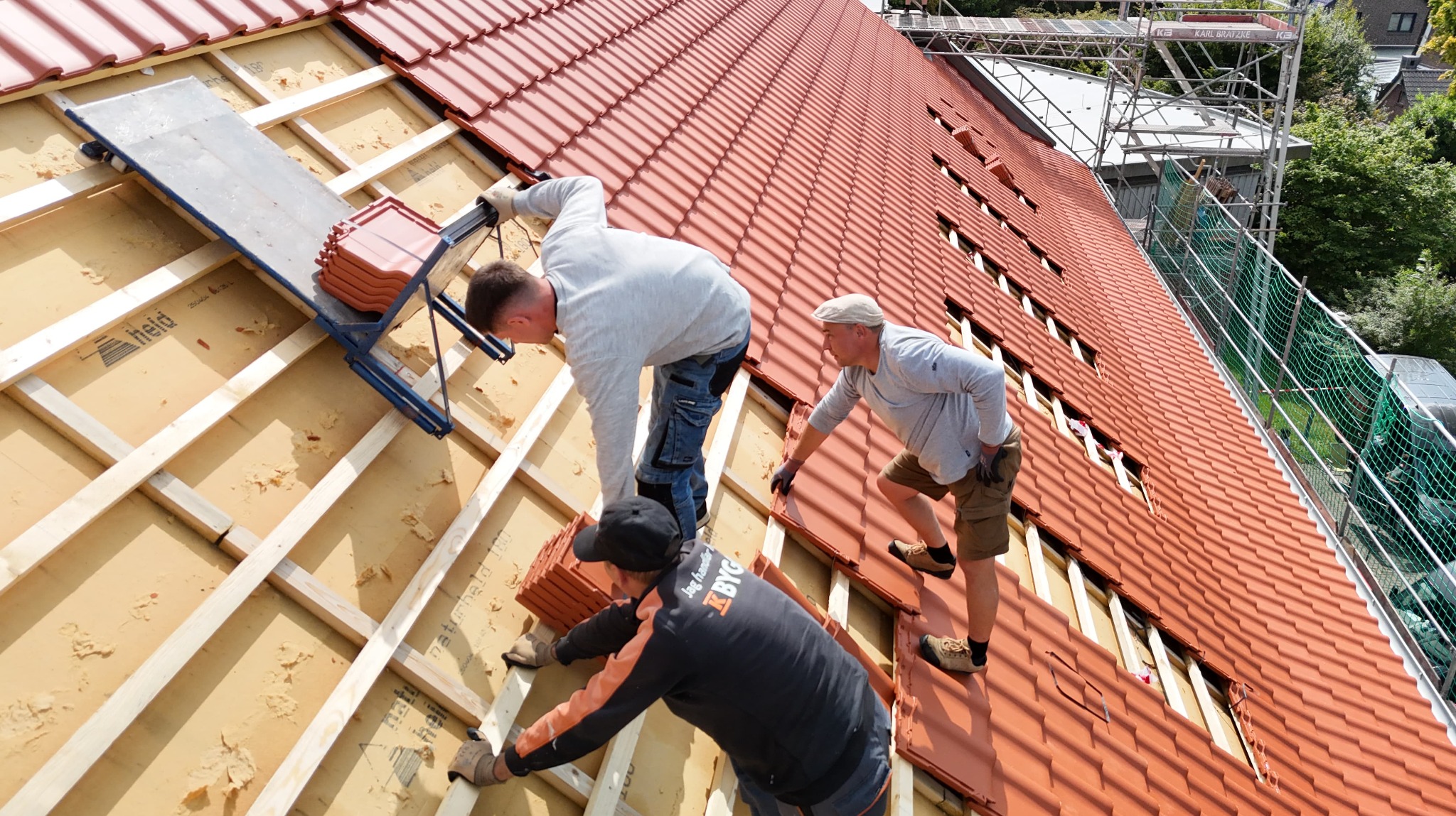 Montaż czerwonej dachówki na ocieplonym poddaszu przez trzech dekarzy. Widoczne rusztowania i narzędzia. Praca zespołowa na wysokości.