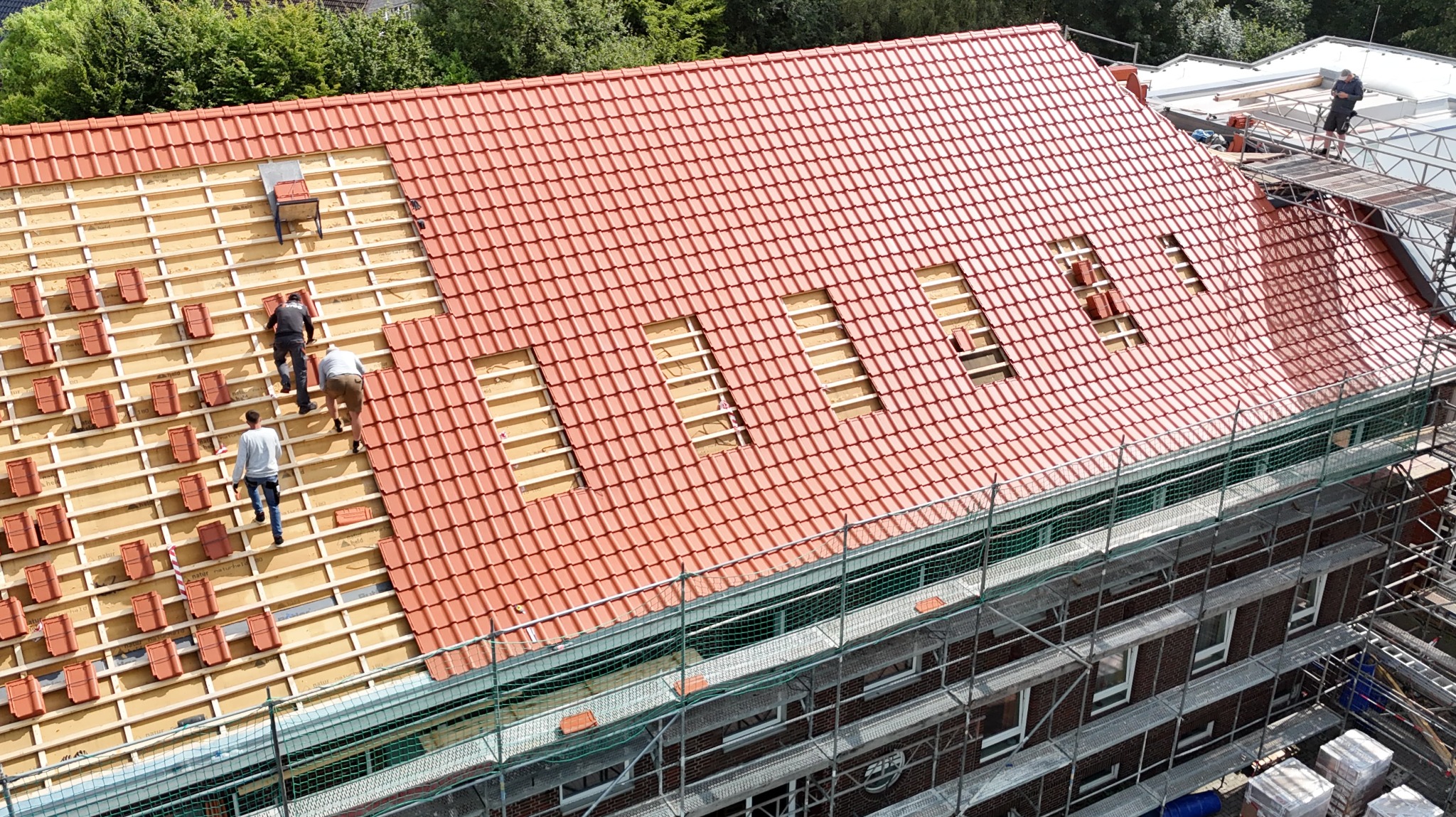 Montaż czerwonej dachówki na drewnianej konstrukcji dachu budynku. Widoczni pracownicy na dachu i rusztowanie przy ścianie budynku. Ujęcie z góry.