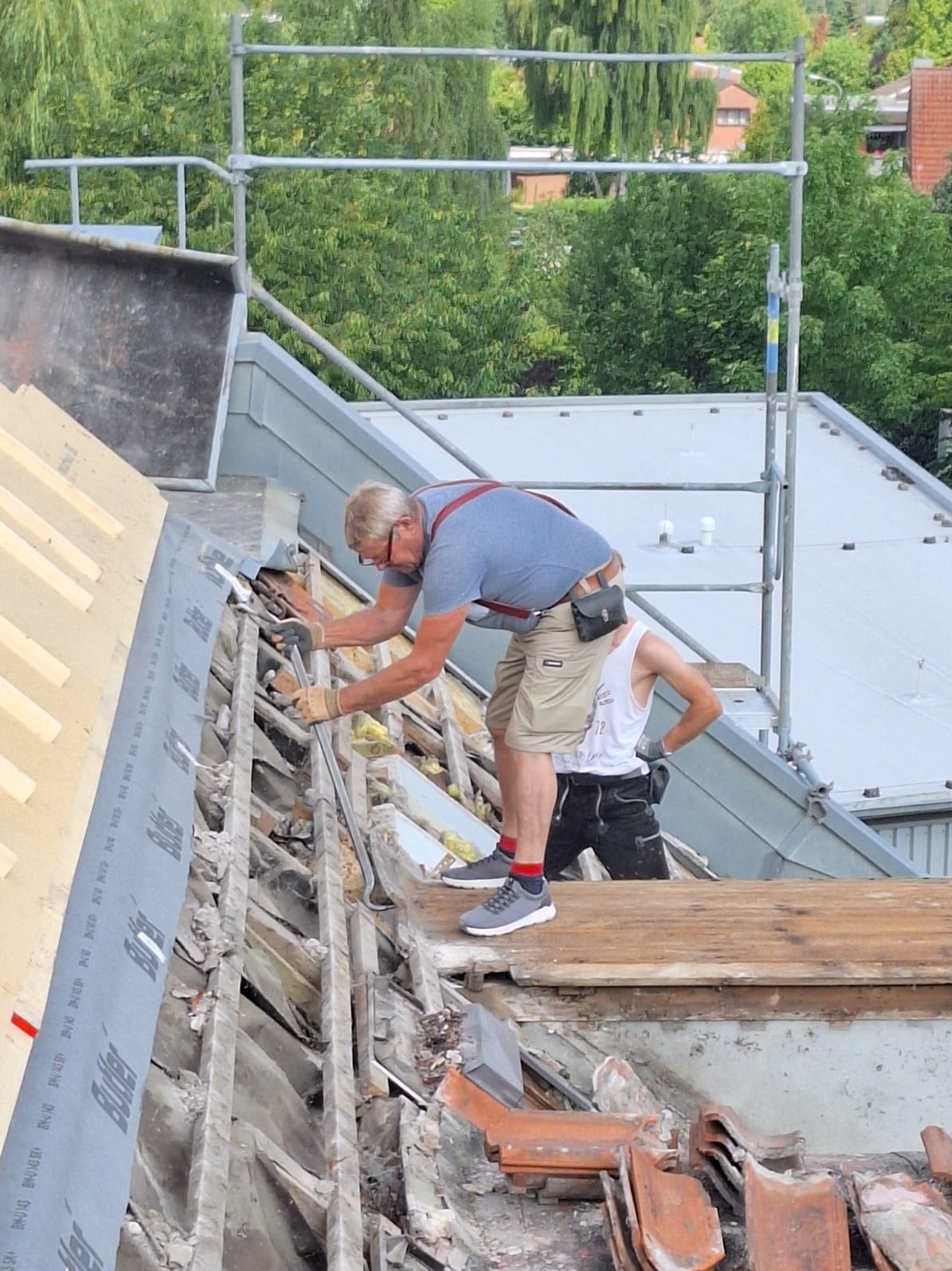 Demontaż starego pokrycia dachowego przez dwóch mężczyzn w okularach, jeden z nich w szelkach, na tle zieleni i rusztowania. Widoczne narzędzia i fragmenty dachówki.
