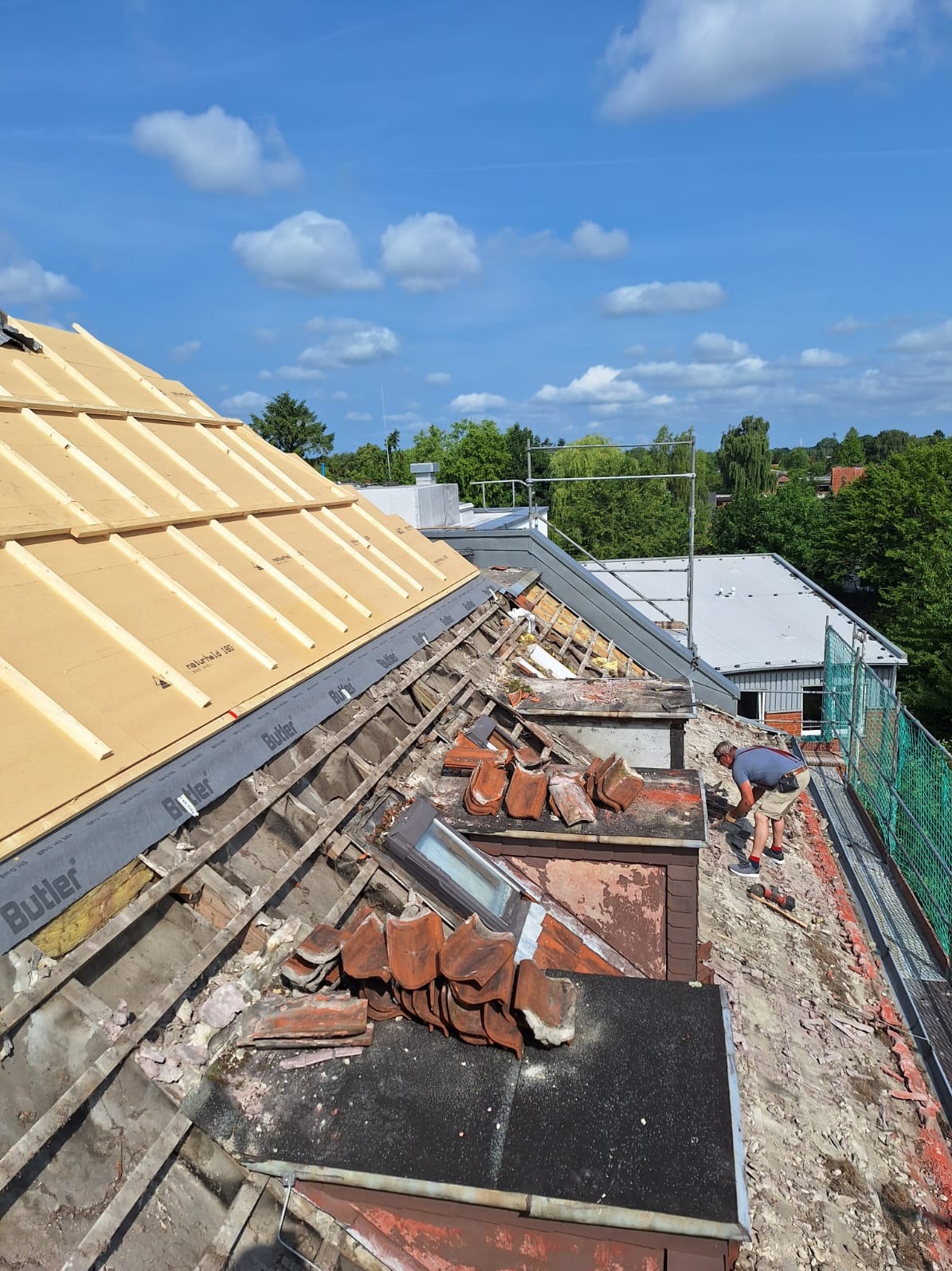 Demontaż starego pokrycia dachowego z widocznymi elementami konstrukcji i oknem dachowym. Prace dekarskie w toku, z fragmentami nowego ocieplenia. Fachowiec przy pracy.