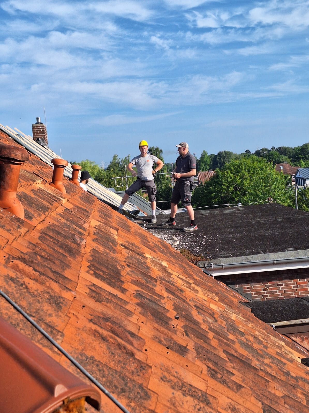 Dwóch dekarzy na dachu z czerwonej dachówki, jeden w żółtym kasku, drugi z papierosem. W tle błękitne niebo i zieleń drzew. Częściowo zdjęta dachówka.