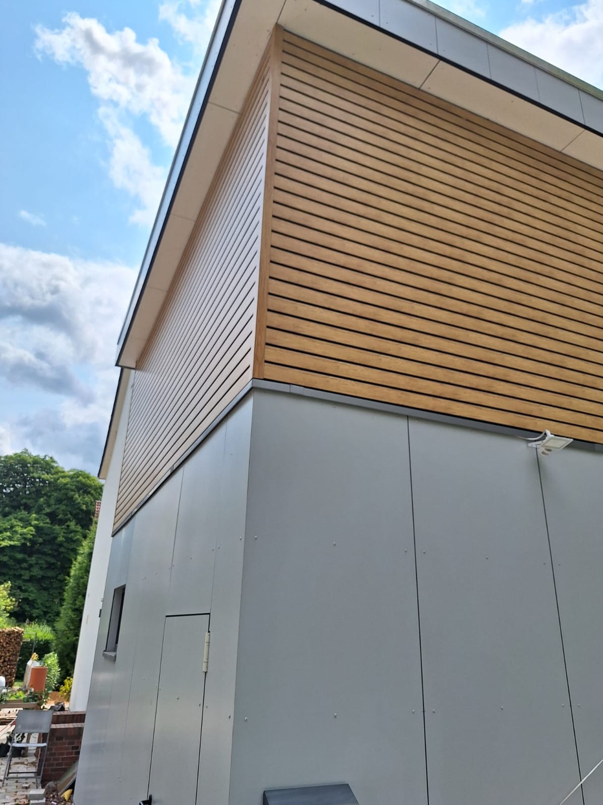 Elewacja budynku z szarymi panelami i drewnianą okładziną. Widoczny fragment dachu, okno i drzwi. Ujęcie z dołu, podkreślające nowoczesny design i materiały wykończeniowe.