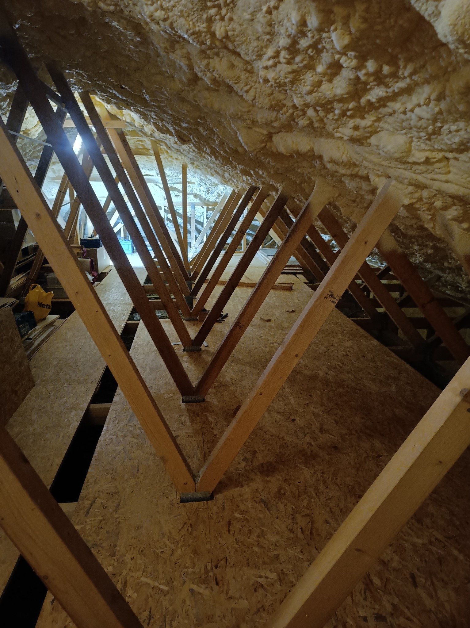 Skośny strych z drewnianą konstrukcją i piankową izolacją. Widoczne deski podłogowe i elementy więźby dachowej. Oświetlenie w tle.