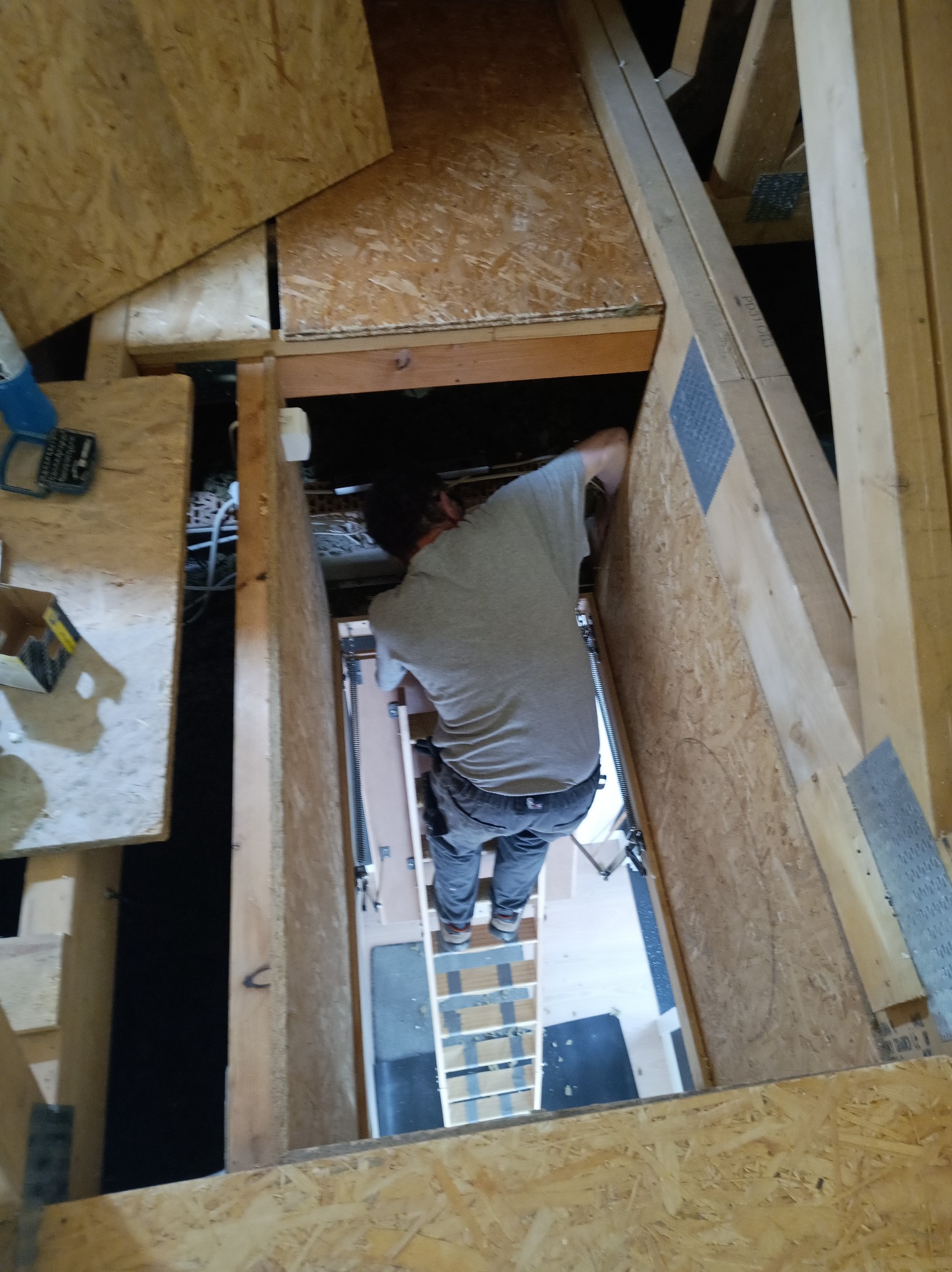 Pracownik montuje płytę OSB w przestrzeni poddasza, widoczna drabina i elementy konstrukcyjne dachu. Ujęcie z góry, podkreślające trudność dostępu.