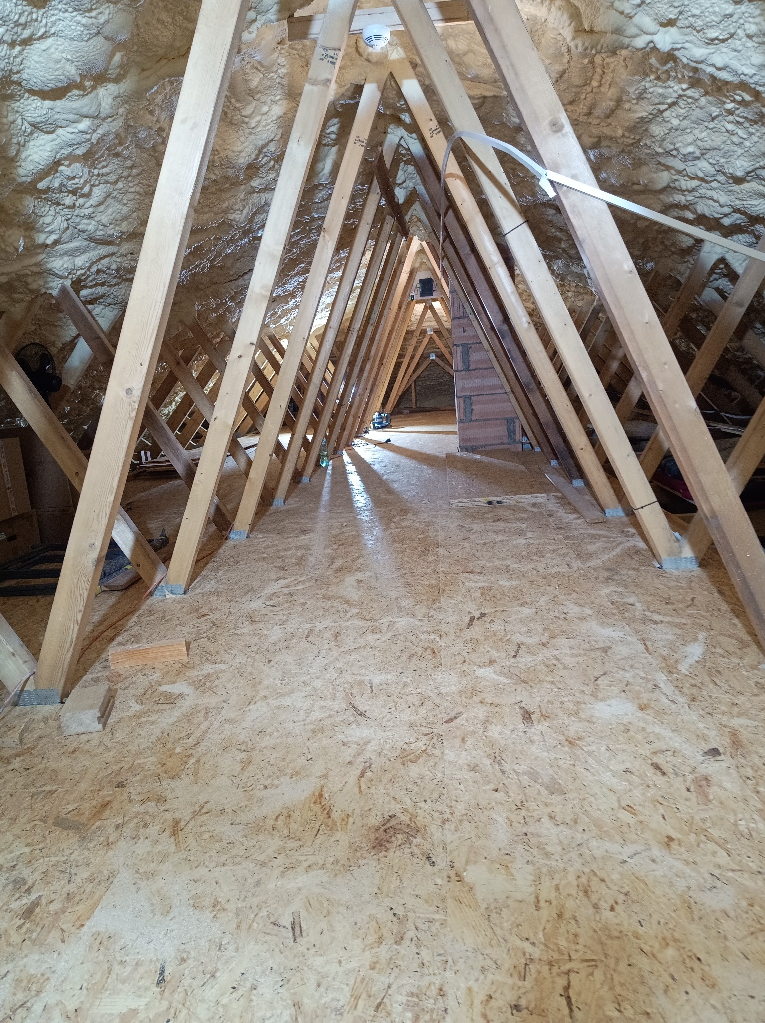 Szkielet drewniany poddasza z widoczną izolacją piankową na skośnych ścianach. Perspektywa z podłogi, ukazująca konstrukcję więźby dachowej i przygotowanie do dalszych prac.