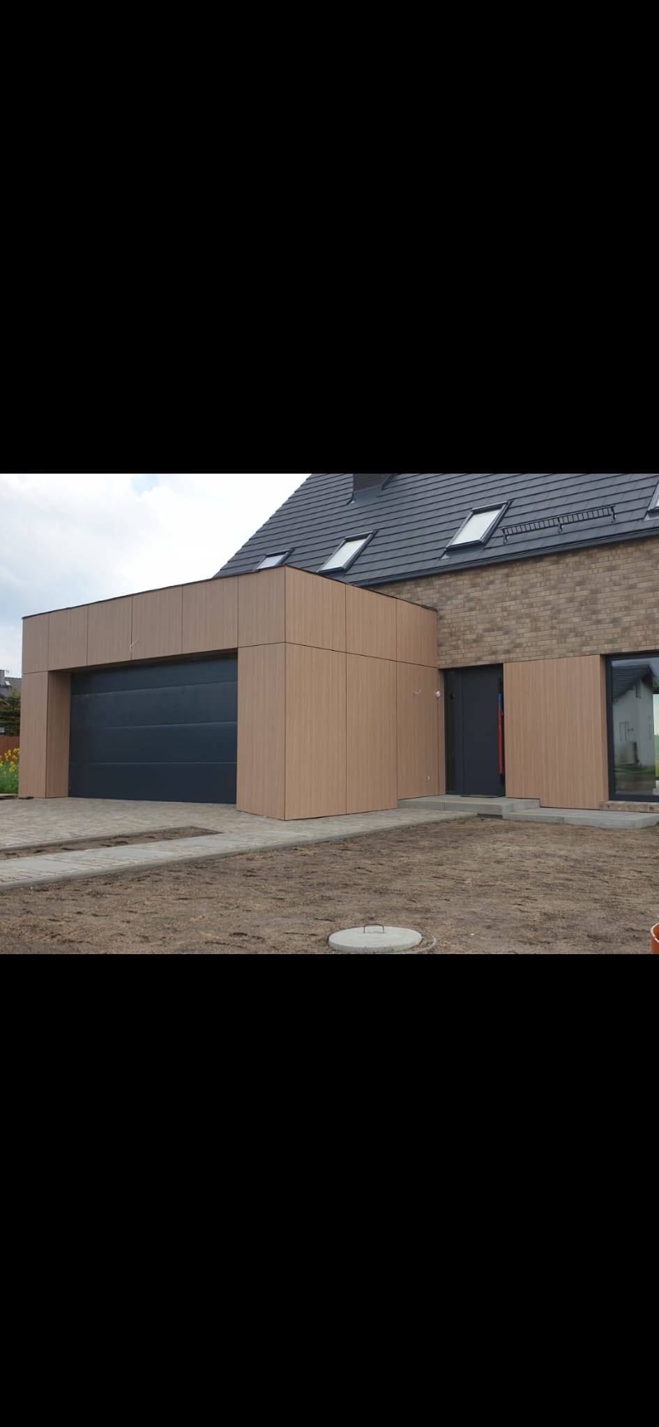 Nowoczesny dom z elewacją z paneli drewnopodobnych i grafitową bramą garażową. Widoczne okna dachowe i ceglana ściana. Całość w stonowanej kolorystyce, na tle nieba.