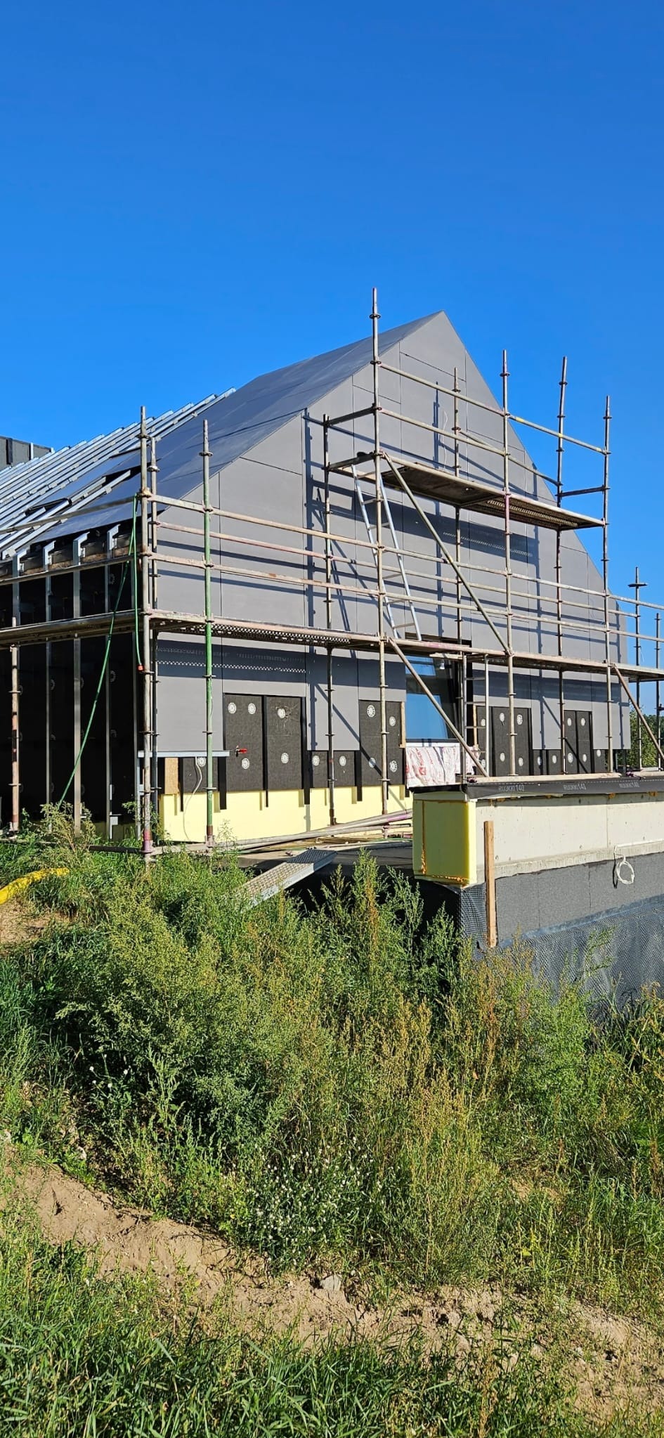 Dom w trakcie ocieplania: szare panele elewacyjne, rusztowania, widoczne warstwy izolacji. Budynek otoczony zielenią, błękitne niebo w tle.