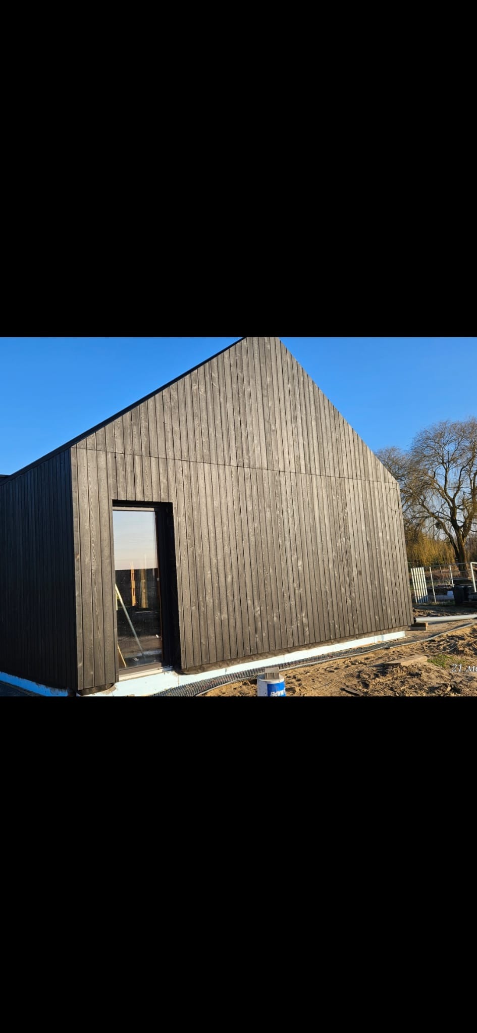 Nowoczesny dom jednorodzinny z elewacją z ciemnego drewna, widok z zewnątrz, błękitne niebo i drzewo w tle, okno z odbiciem krajobrazu, minimalistyczny design.