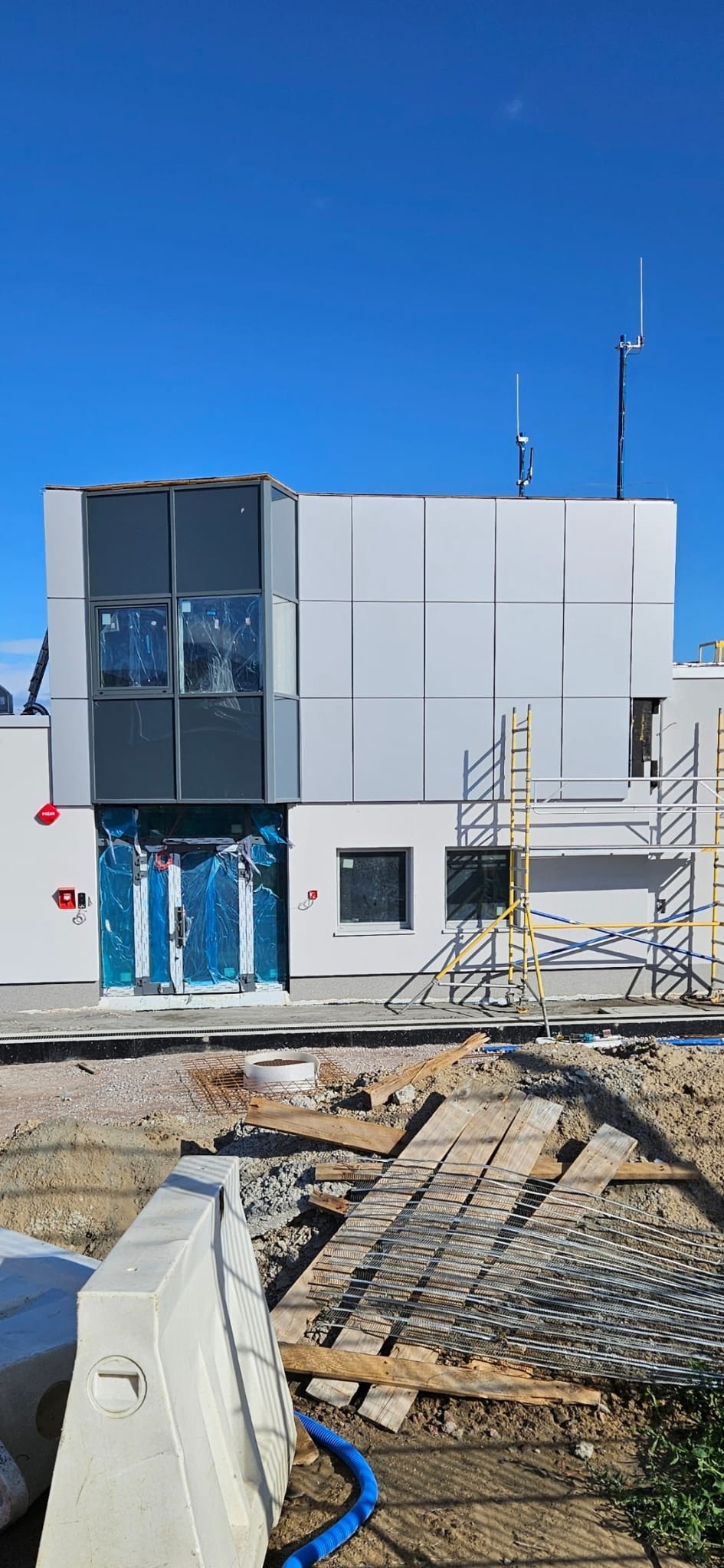 Budynek w trakcie prac elewacyjnych. Widoczne rusztowania, zabezpieczone folią wejście i fragment terenu budowy. Elewacja częściowo wykończona panelami.