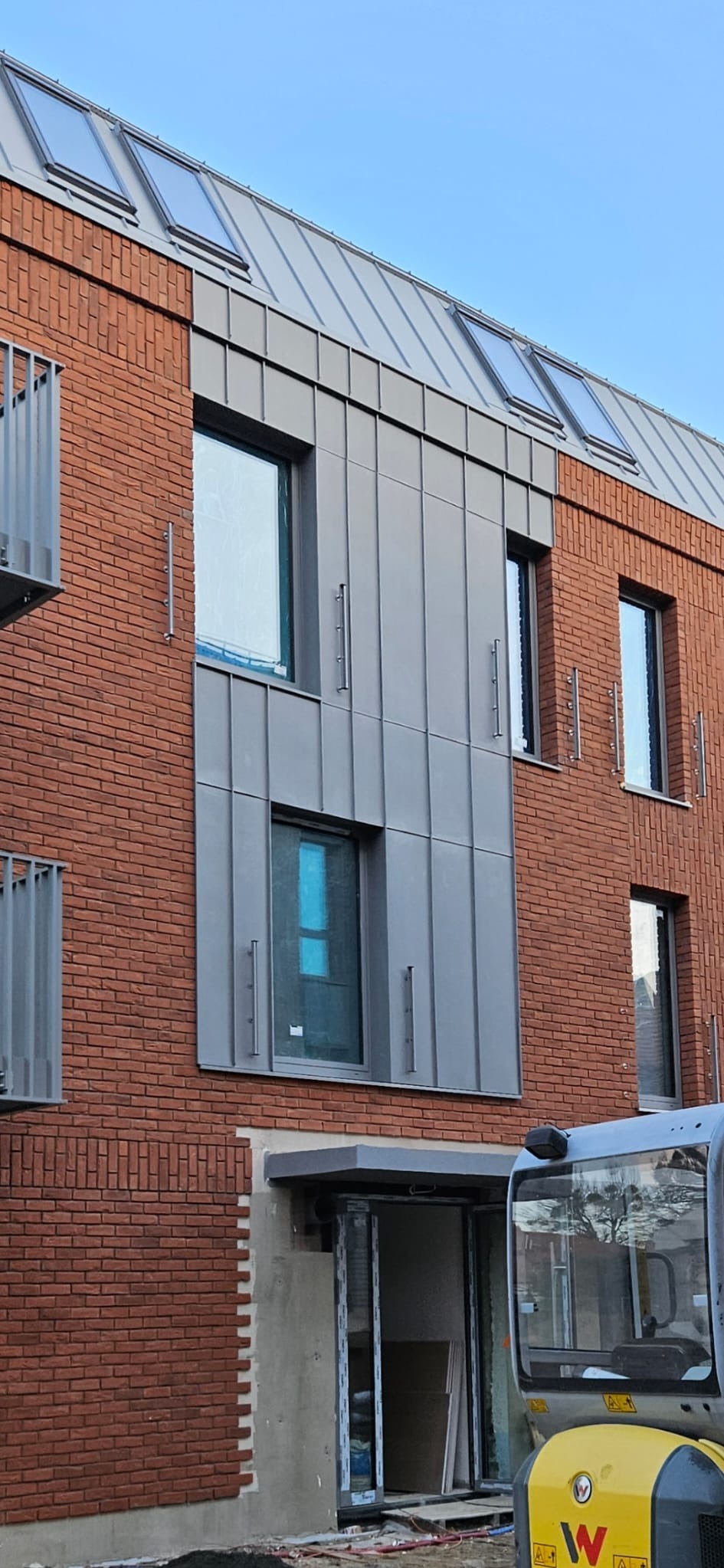Ceglana elewacja budynku z szarymi panelami i metalowym dachem. Widoczne okna i balkon. Częściowo zasłonięte wejście i maszyna budowlana.