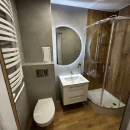 Nowoczesna łazienka z kabiną prysznicową, umywalką z podświetlanym lustrem i grzejnikiem drabinkowym. Płytki imitujące drewno na podłodze i ścianie kabiny.