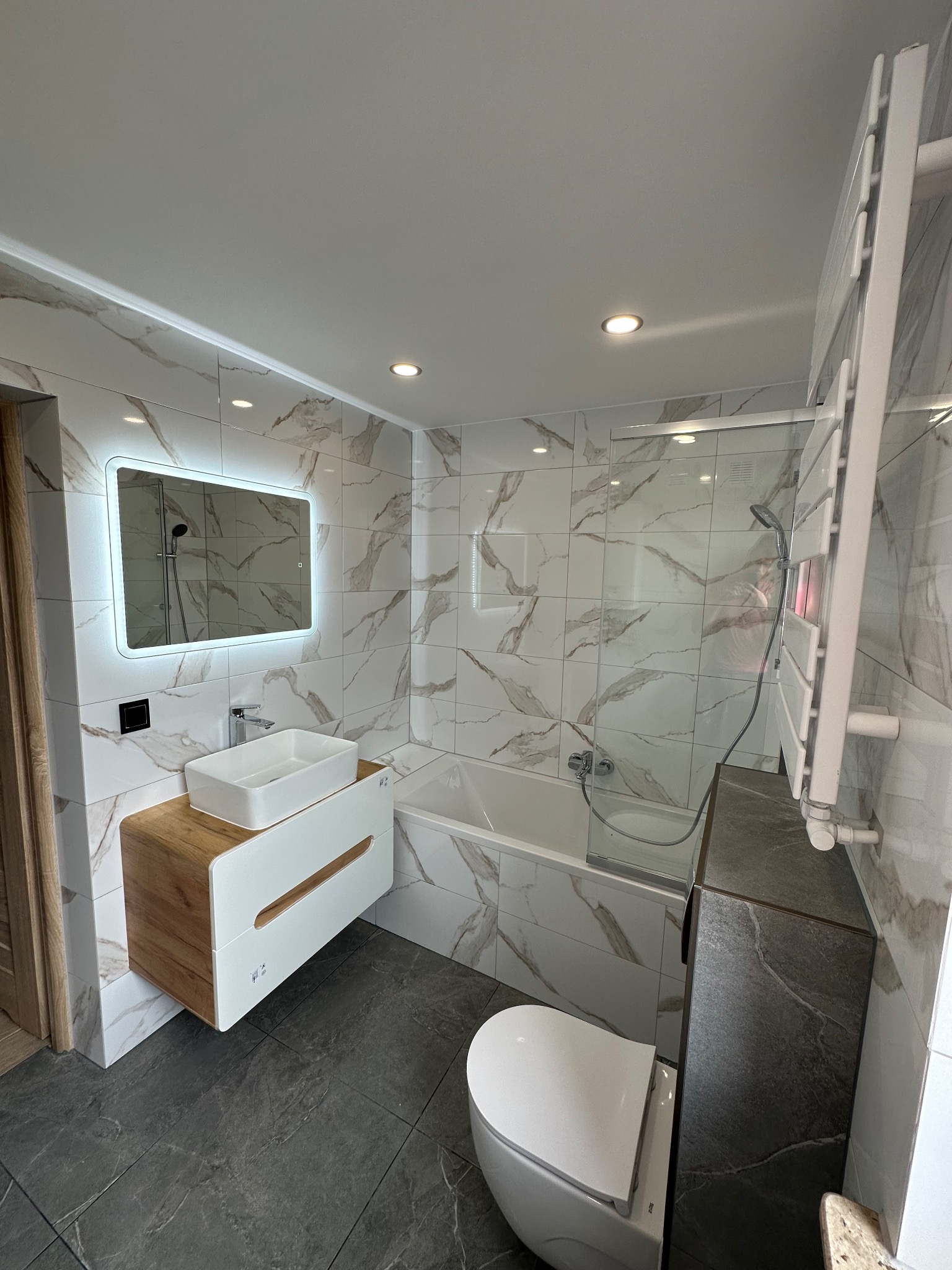 Nowoczesna łazienka z wanną za szklaną ścianką, umywalką na szafce z drewnianym akcentem i podświetlanym lustrem. Płytki imitujące marmur na ścianach i ciemna podłoga.