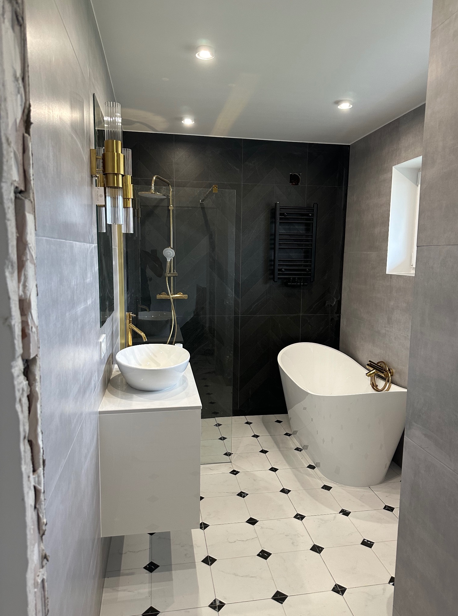 Nowoczesna łazienka po remoncie: biała wanna i umywalka na tle czarnych płytek. Złote akcenty w armaturze i oświetleniu, wzorzysta podłoga.