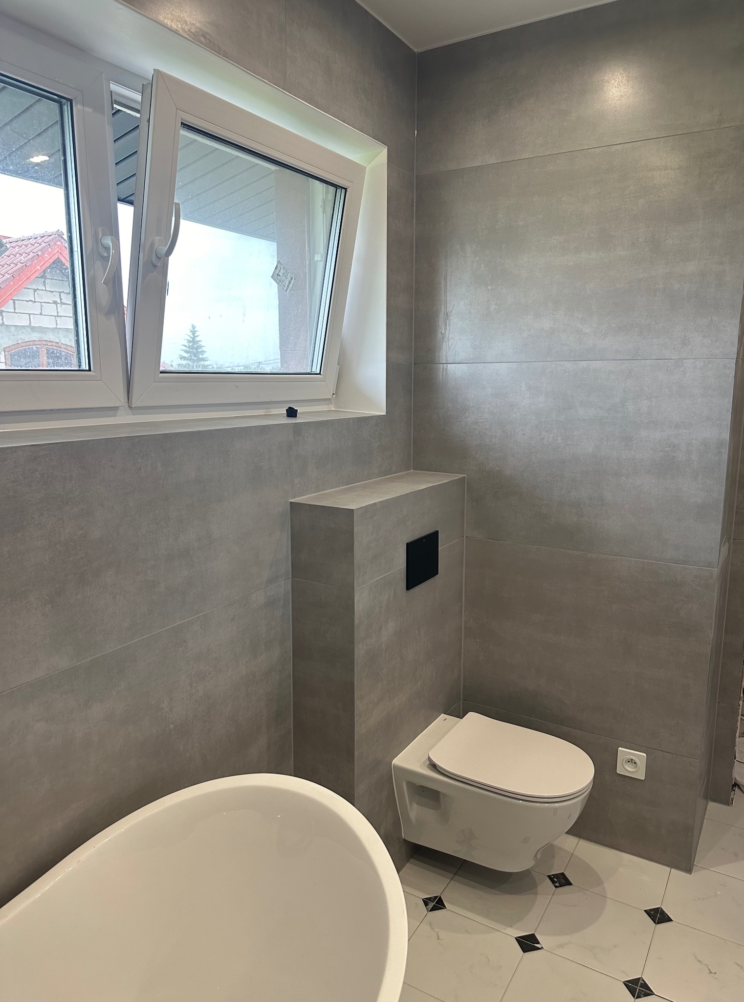 Wykończona łazienka w szarościach z podwieszaną toaletą i oknem w Sanoku. Widoczna wanna i płytki imitujące beton. Nowoczesny design i dbałość o detale.