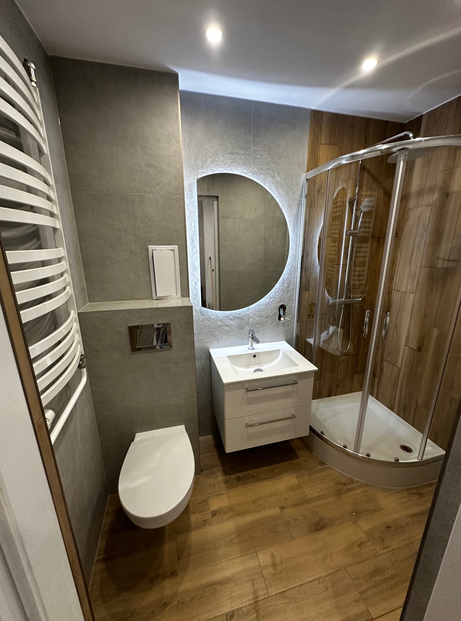 Nowoczesna łazienka z kabiną prysznicową, umywalką z podświetlanym lustrem i grzejnikiem drabinkowym. Płytki imitujące drewno na podłodze i ścianie kabiny.