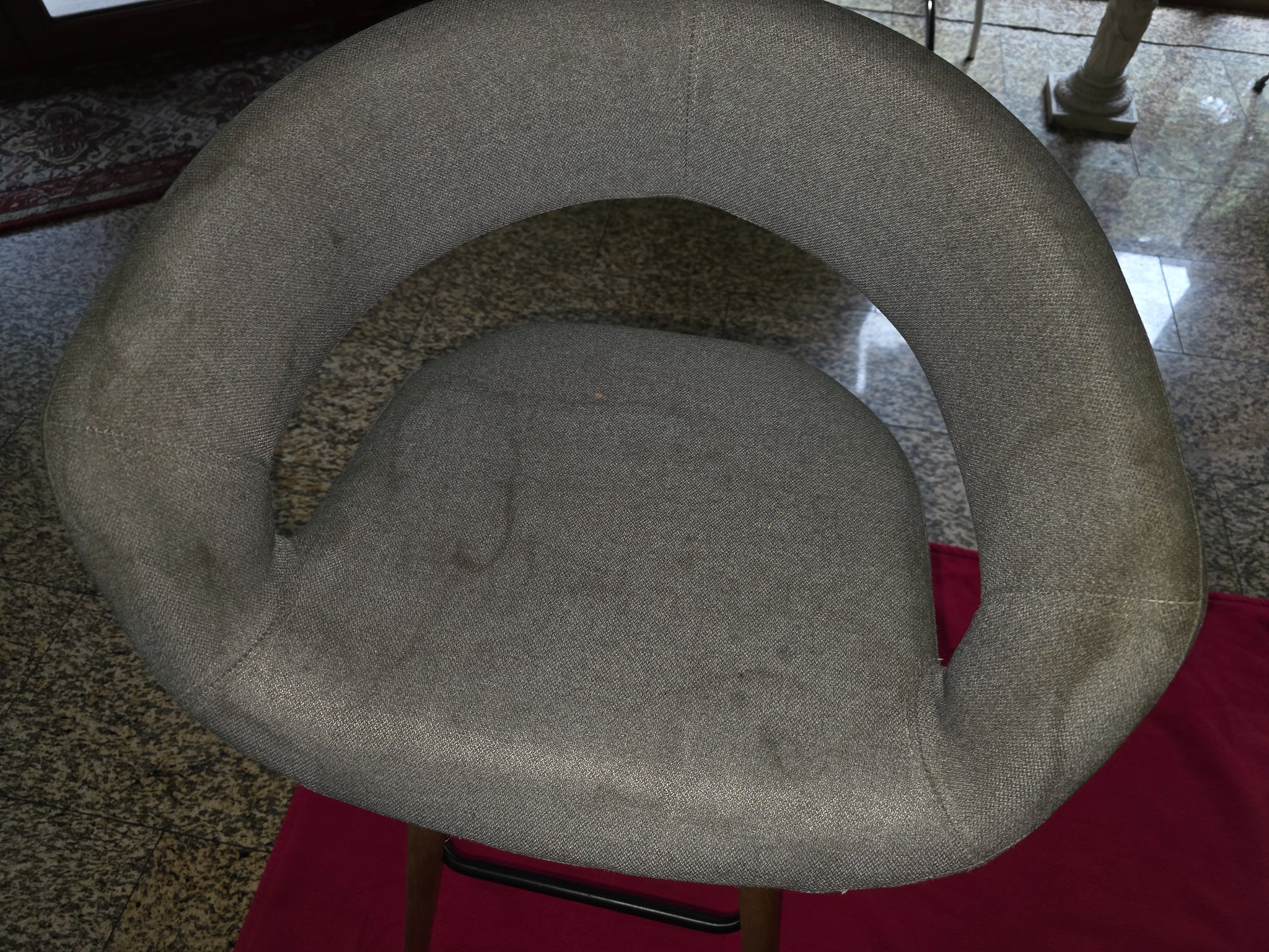 Szare krzesło tapicerowane z widocznymi plamami na siedzisku i oparciu, ukazane z góry na tle marmurowej podłogi i fragmentu czerwonego materiału.