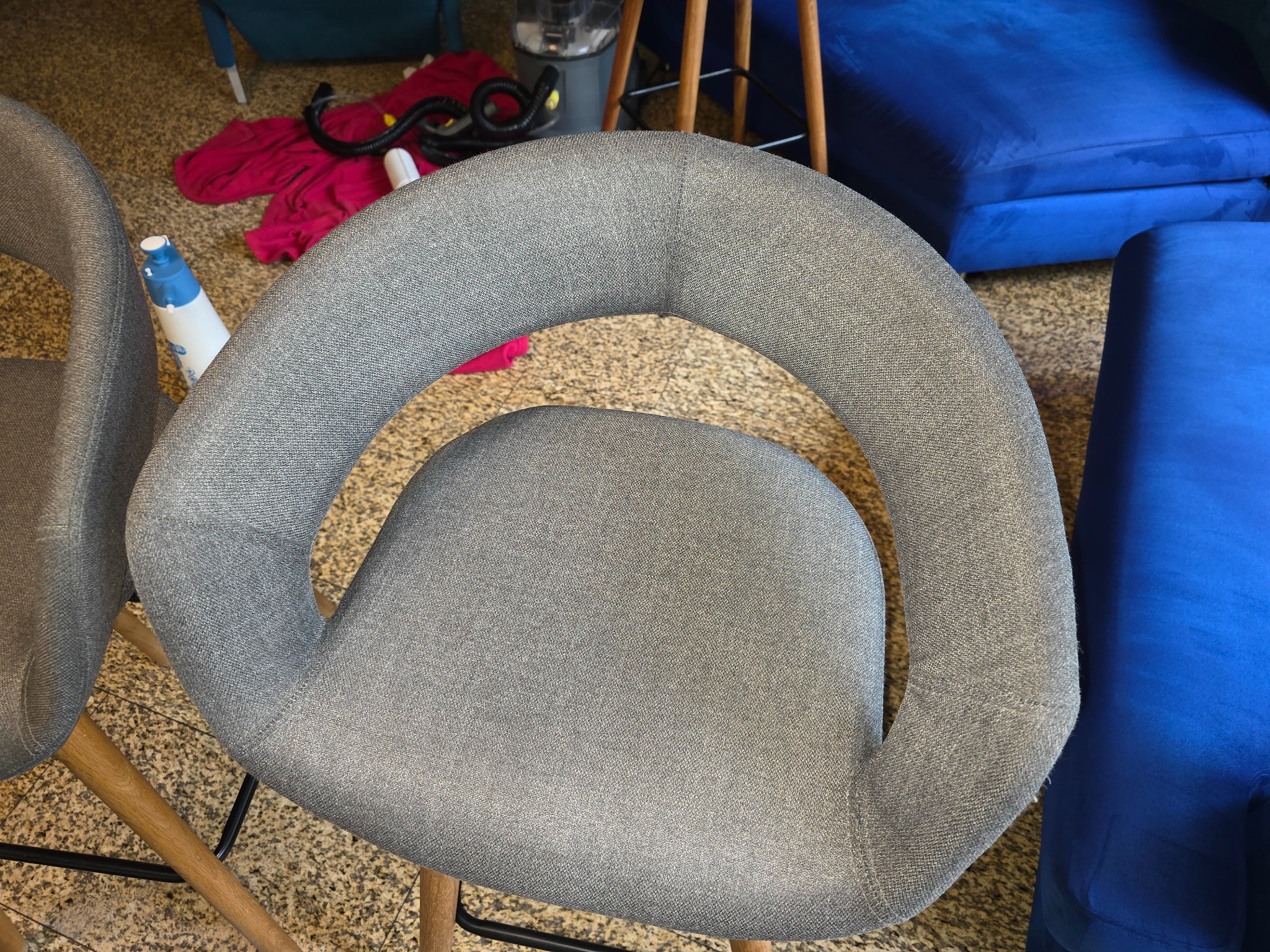 Szare krzesło tapicerowane z drewnianymi nogami, widoczne obok niebieskiej kanapy i sprzętu do czyszczenia, sugerujące prace porządkowe lub pielęgnację mebli.