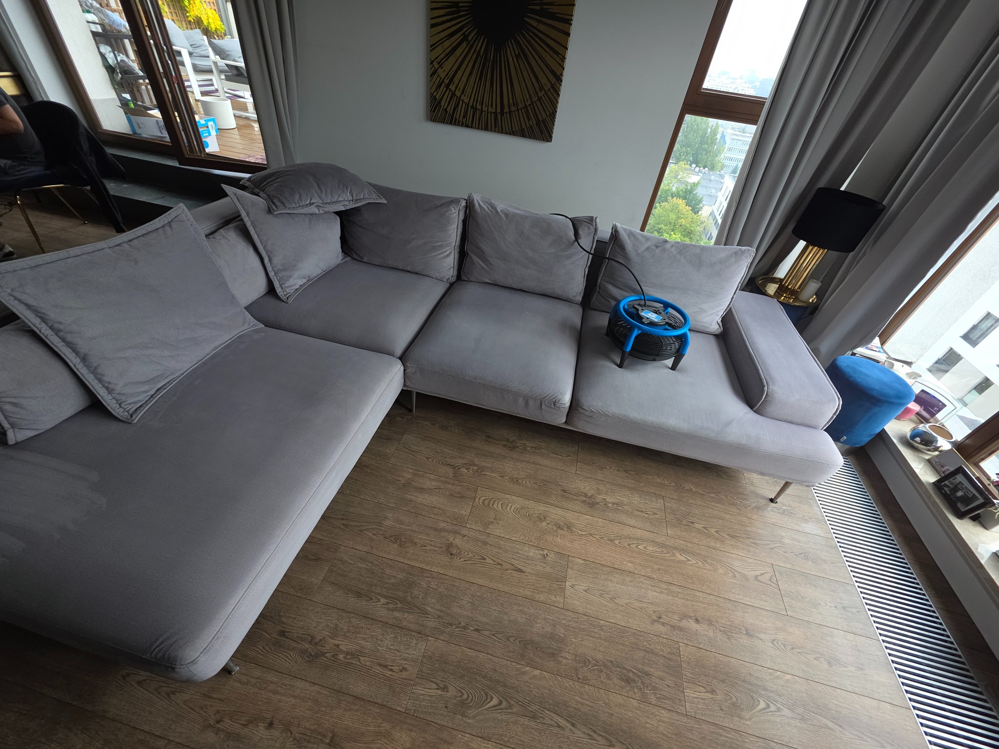 Szara, modułowa kanapa narożna z poduszkami, na której stoi niebieski wentylator do prania tapicerki. W tle okno z widokiem na miasto i designerska lampa.
