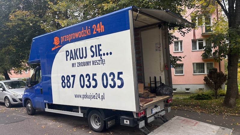 Niebiesko-biała furgonetka z otwartą przestrzenią ładunkową i napisem 'przeprowadzki 24h' na tle bloku mieszkalnego i drzew.
