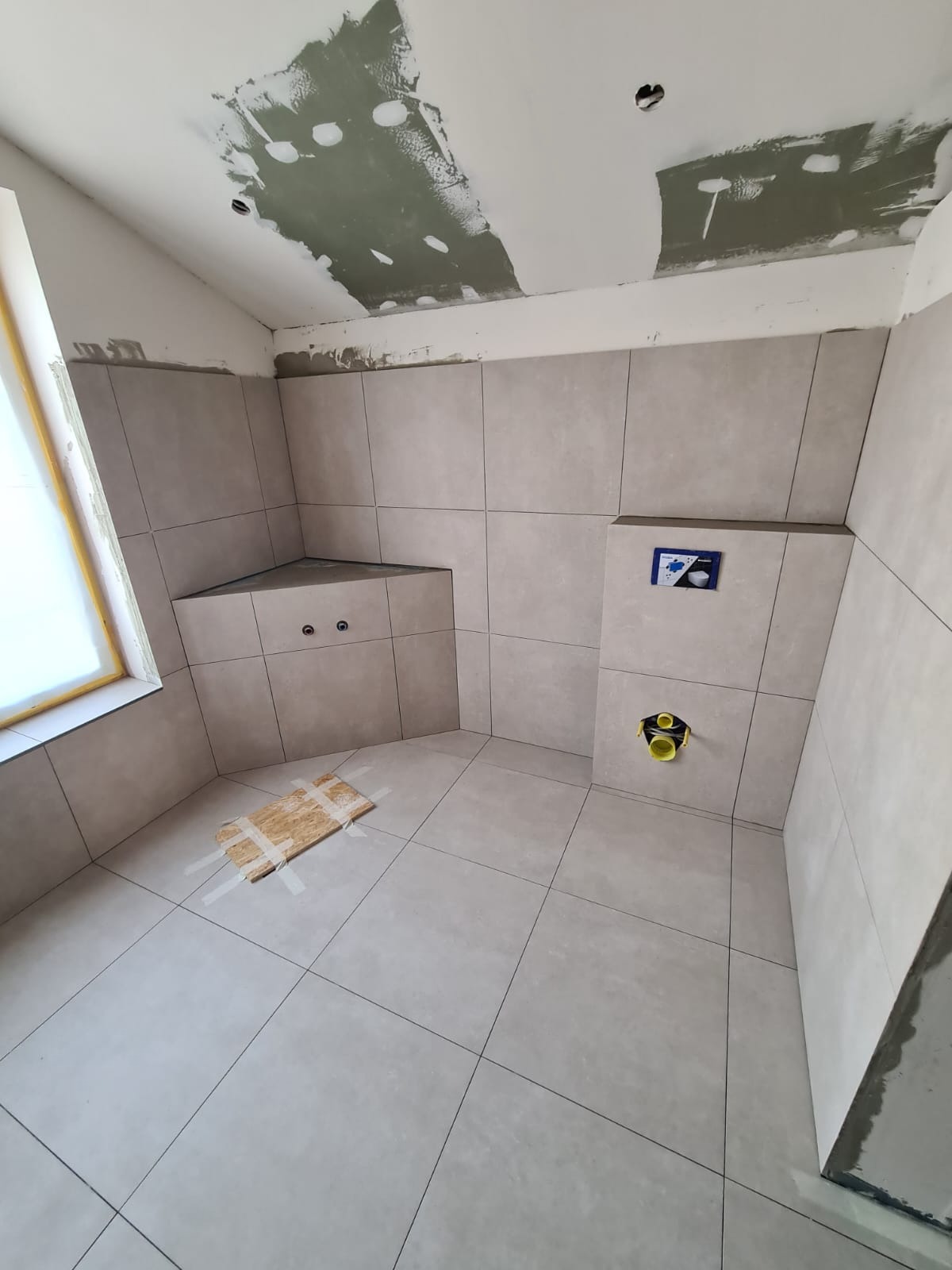 Wykończona łazienka z beżowymi płytkami, widoczne elementy instalacji podtynkowej i fragment sufitu w trakcie prac wykończeniowych. Na podłodze leży zabezpieczenie.