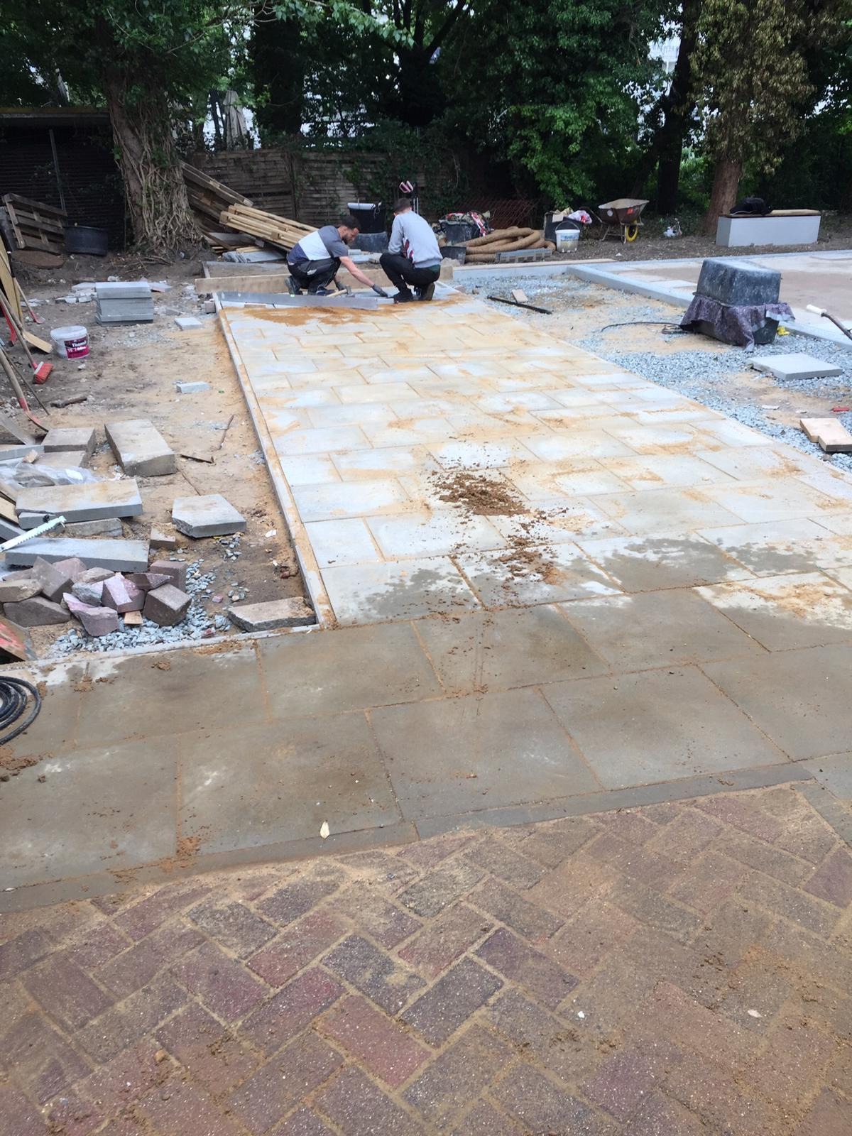 Układanie jasnoszarej kostki brukowej na posesji, widoczni dwaj pracownicy w trakcie montażu. Częściowo ułożona nawierzchnia z kostki w otoczeniu zieleni.