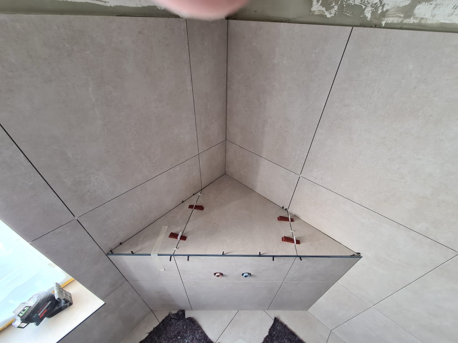 Montaż jasnych, kwadratowych płytek w rogu pomieszczenia. Widoczne narzędzia glazurnicze i elementy mocujące. Płytki ułożone z zachowaniem fug.