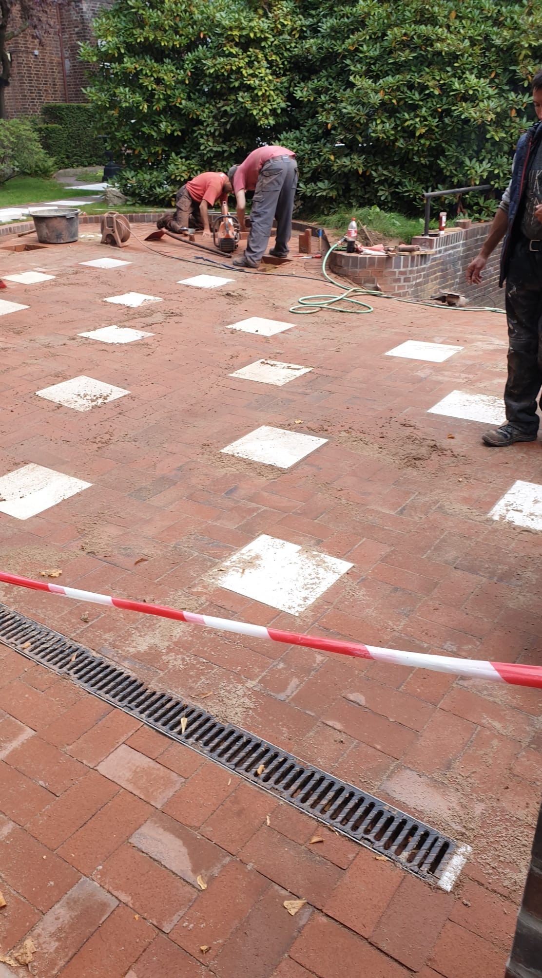 Układanie kostki brukowej z wyznaczonymi miejscami na płyty, widoczny drenaż, dwóch pracowników w trakcie pracy, taśma ostrzegawcza.