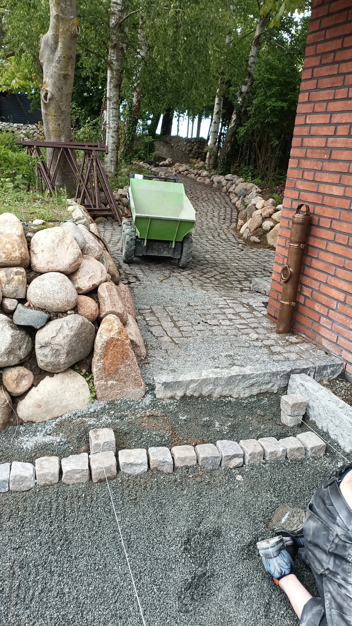 Układanie kostki granitowej: zielona taczka, kamienna ścieżka w ogrodzie, mur z kamieni, ceglany budynek, praca w toku, widok z góry.